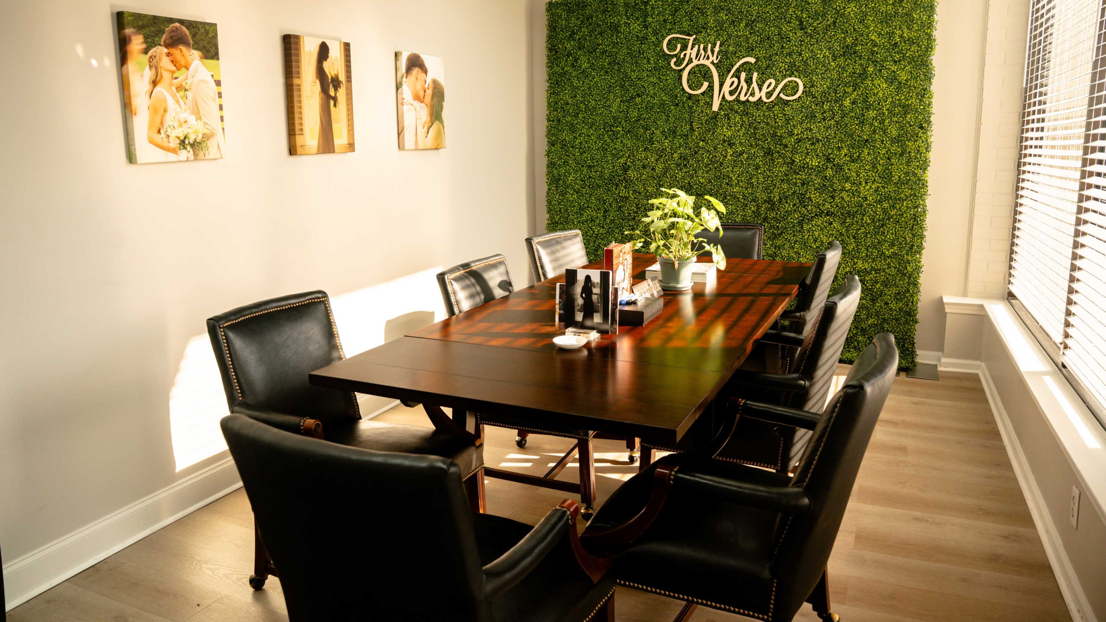 The image shows a conference room with a wooden table surrounded by black chairs, illuminated by natural light, and decorated with framed photos on the walls and a green plant wall with the words "First Verse."