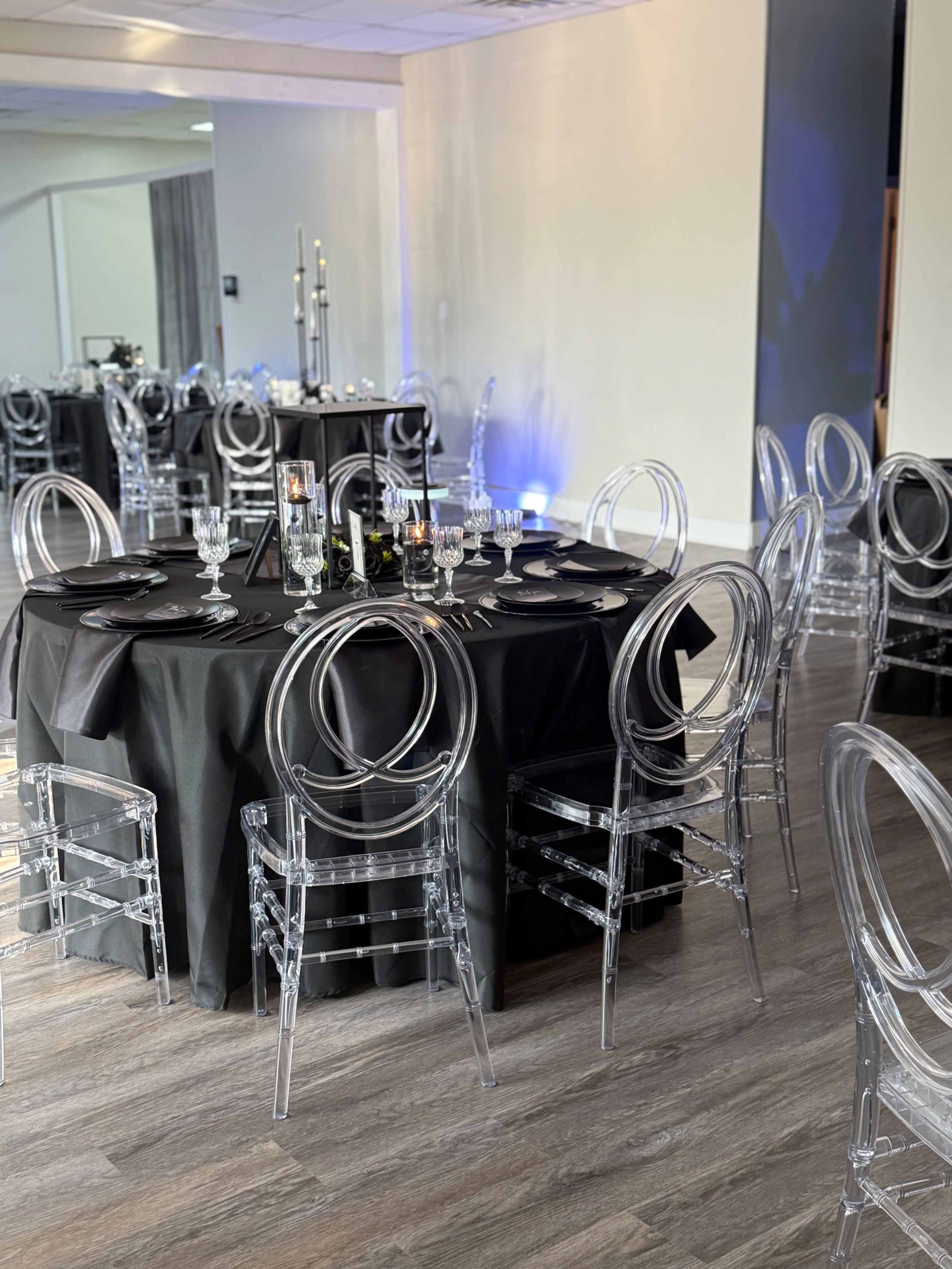 A dining area features round tables covered with black tablecloths and set with clear acrylic chairs, lit by soft ambient lighting.
