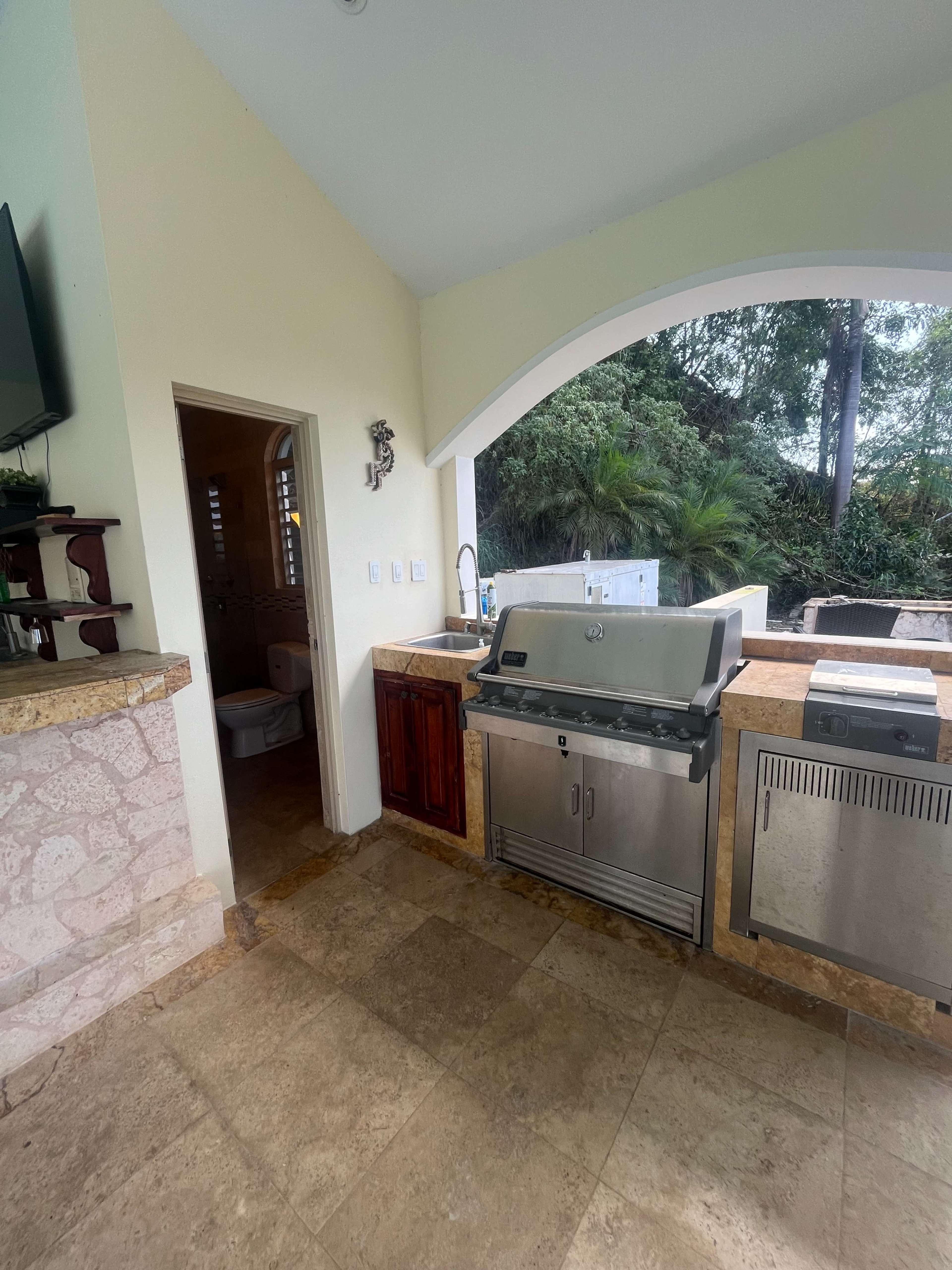 The image shows an outdoor kitchen area featuring a stainless steel grill, a refrigerator, and a view of greenery through a large window.