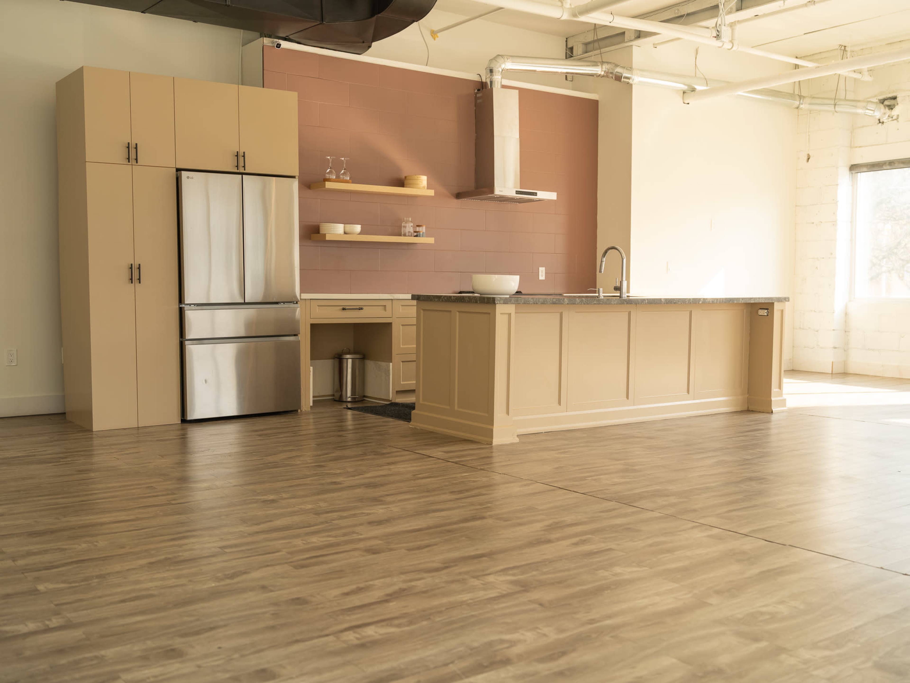 The image shows a modern kitchen with light-colored cabinetry, a stainless steel refrigerator, an island with a countertop, and large windows allowing natural light to fill the space.
