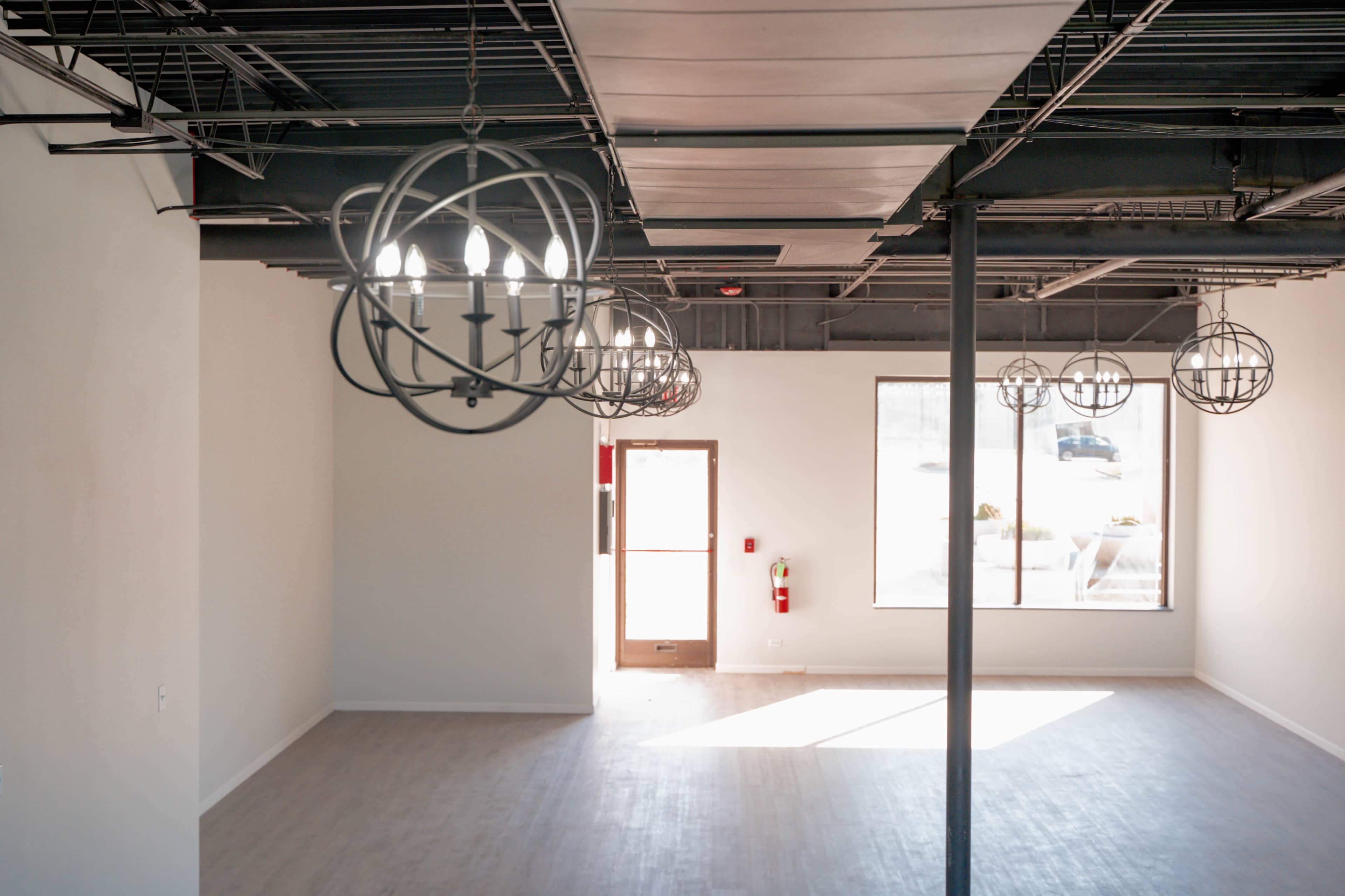 An empty interior space with wooden flooring, a large window, and modern light fixtures hanging from the ceiling.
