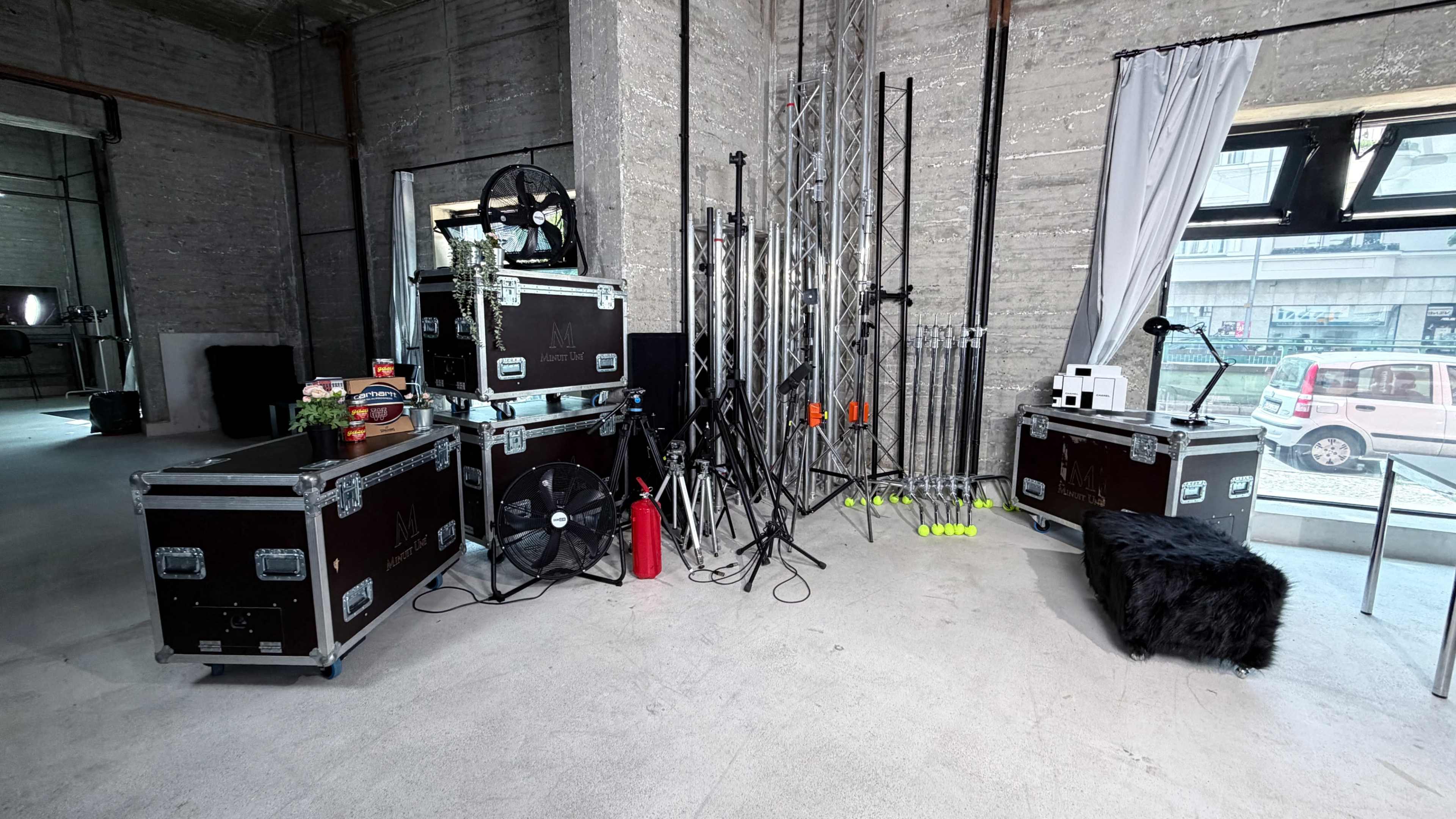 The image shows a workspace with stacked equipment cases, a fan, lighting stands, and a black chair in a minimalist, industrial setting.