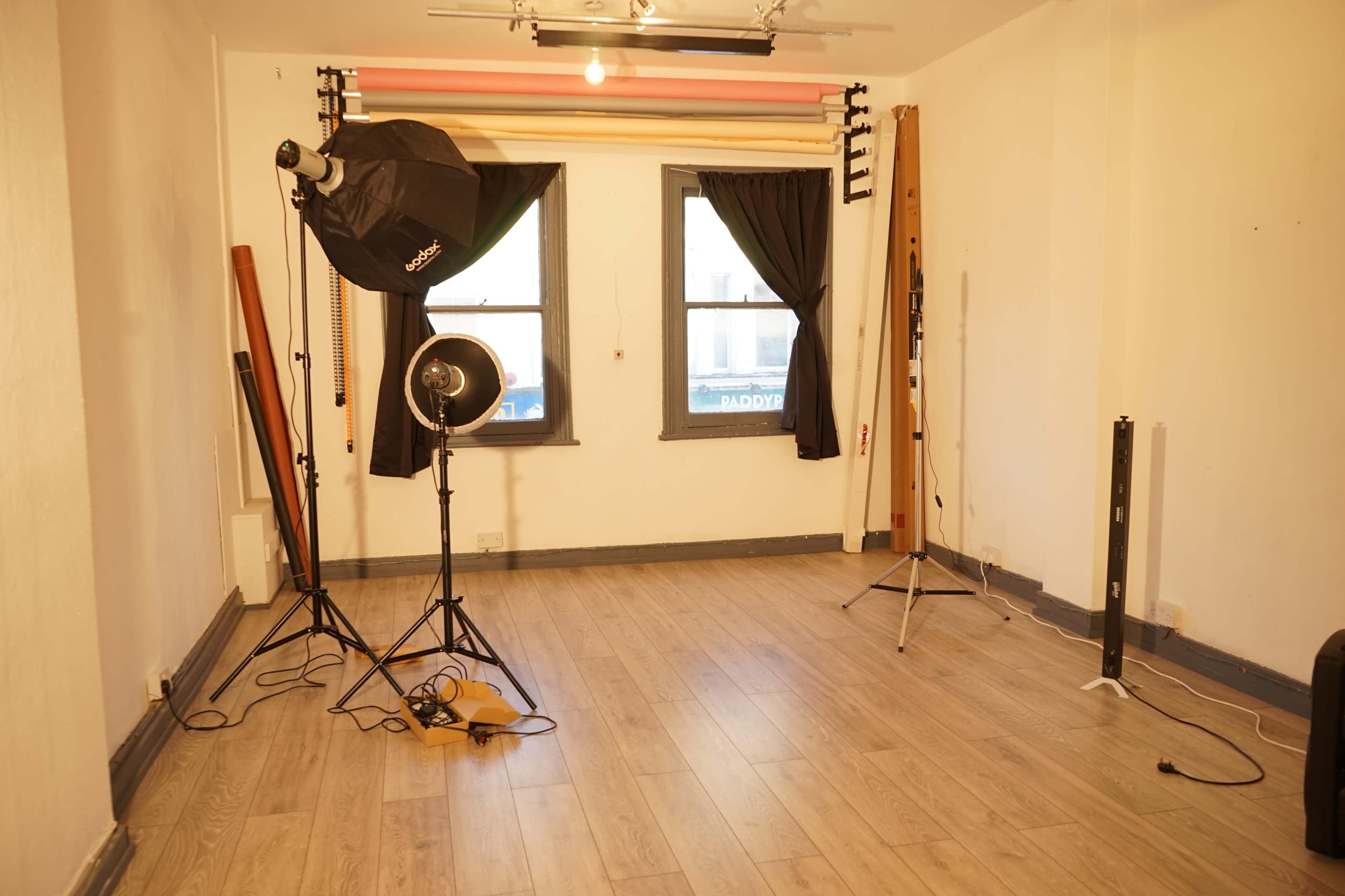 The image shows a well-lit photography studio with various lighting equipment, empty floor space, and two windows dressed with dark curtains.