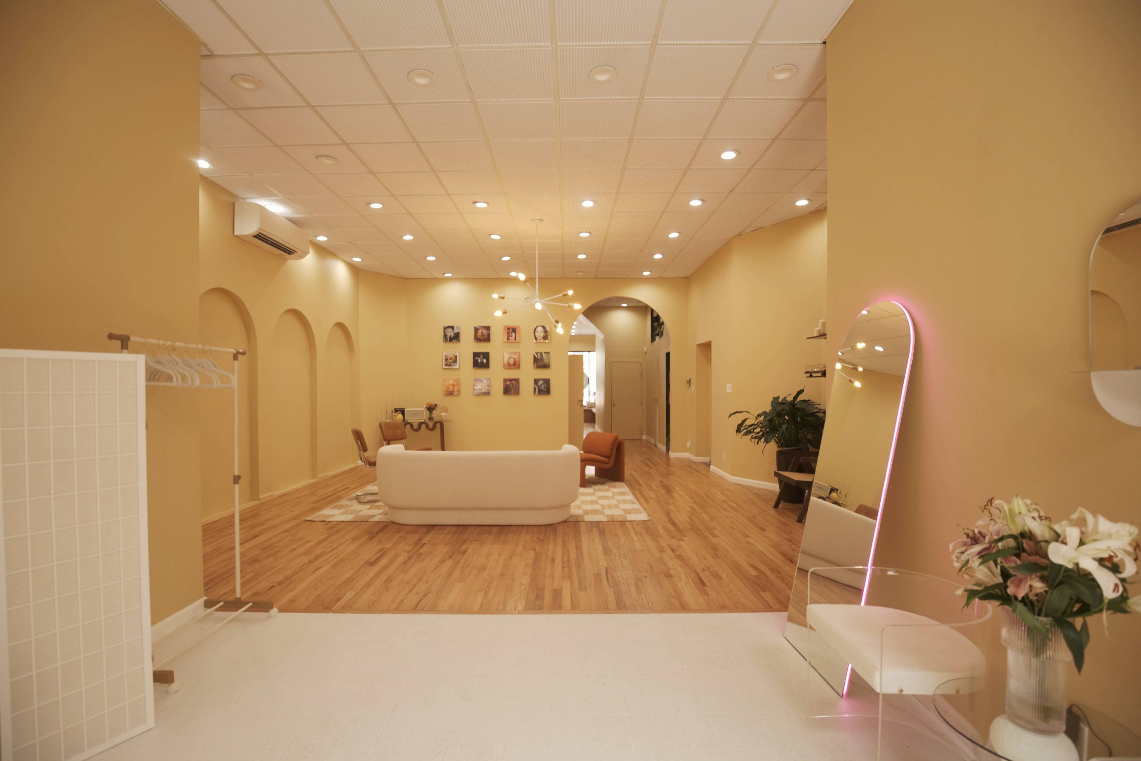 The image shows a spacious, well-lit interior with a light beige color scheme, featuring a seating area, photographs on the walls, and a large mirror on one side.