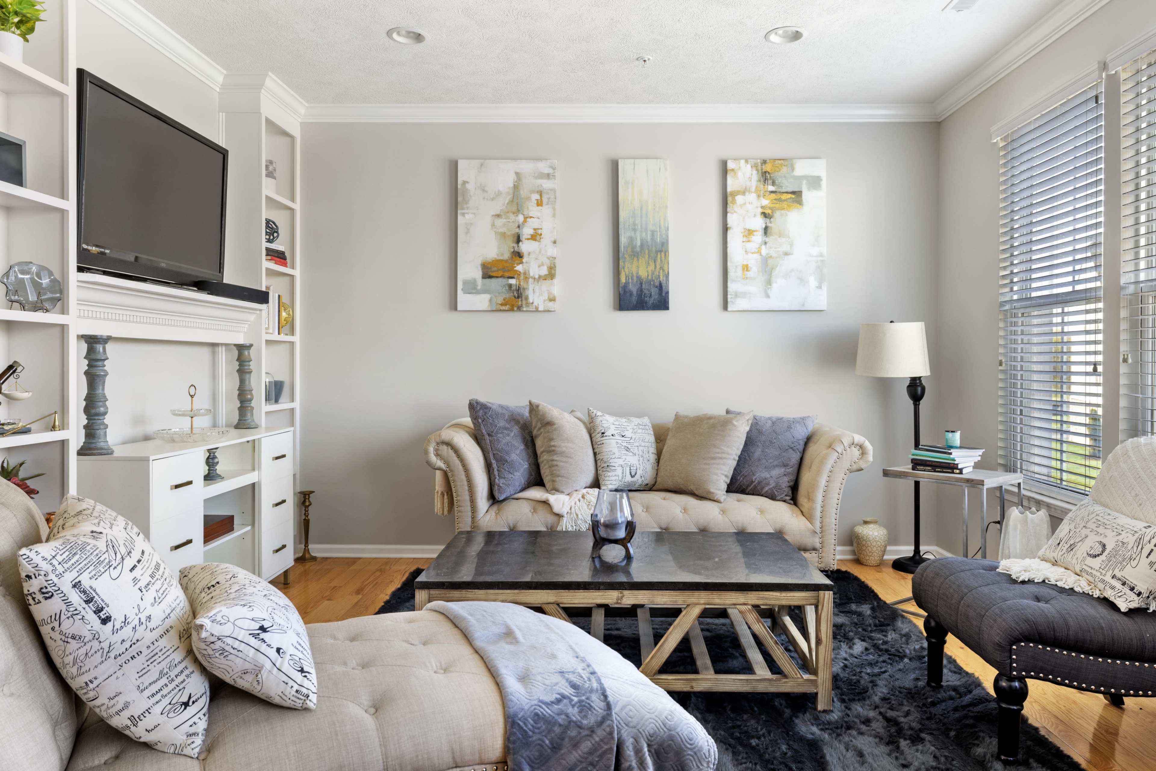 A cozy living room featuring a light-colored sofa, a dark coffee table, two abstract paintings on the wall, and a bright window with blinds.