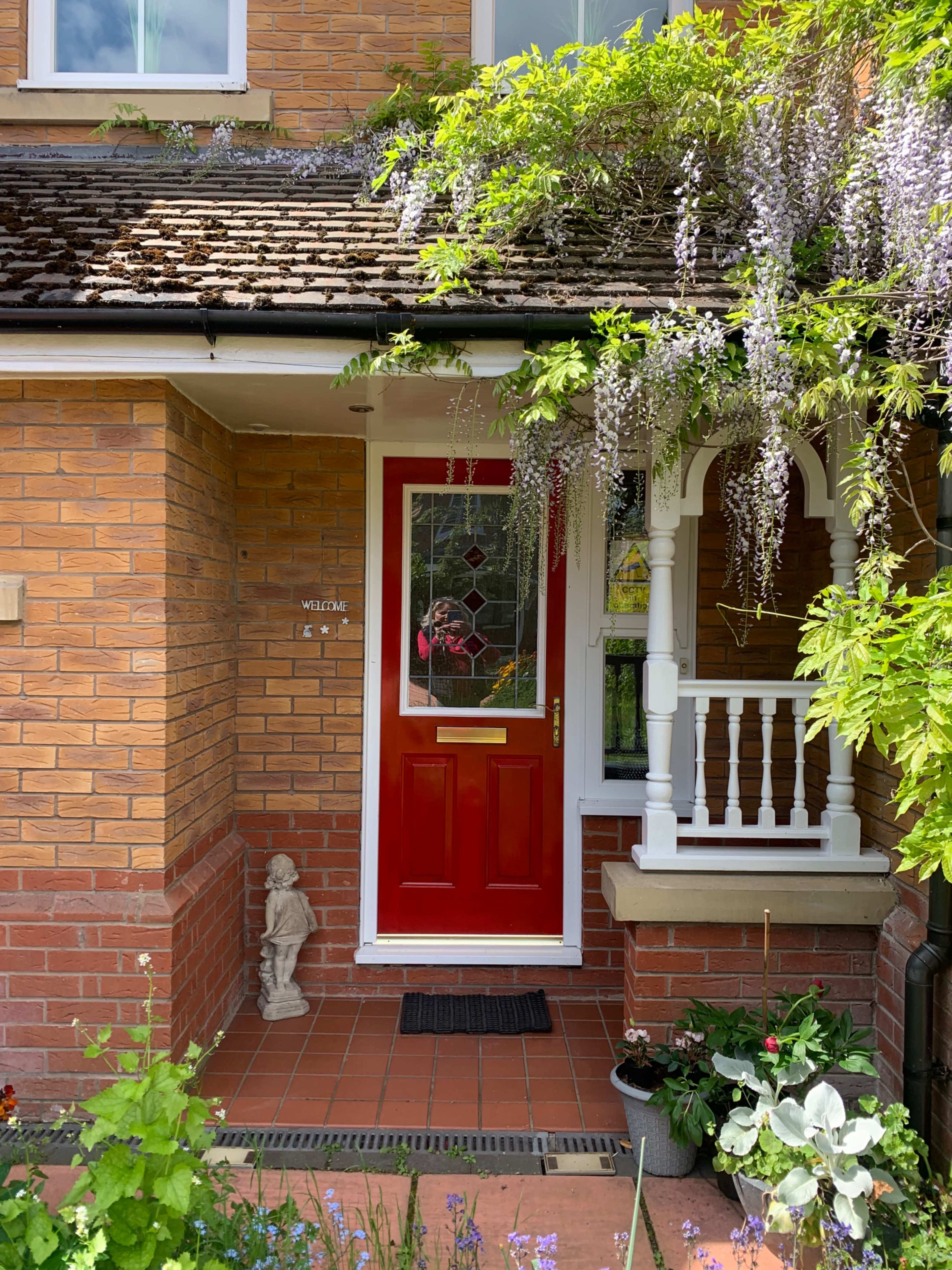 The image shows a brick house entrance with a red front door, a covered porch, hanging wisteria, and a garden with various plants and a decorative statue.