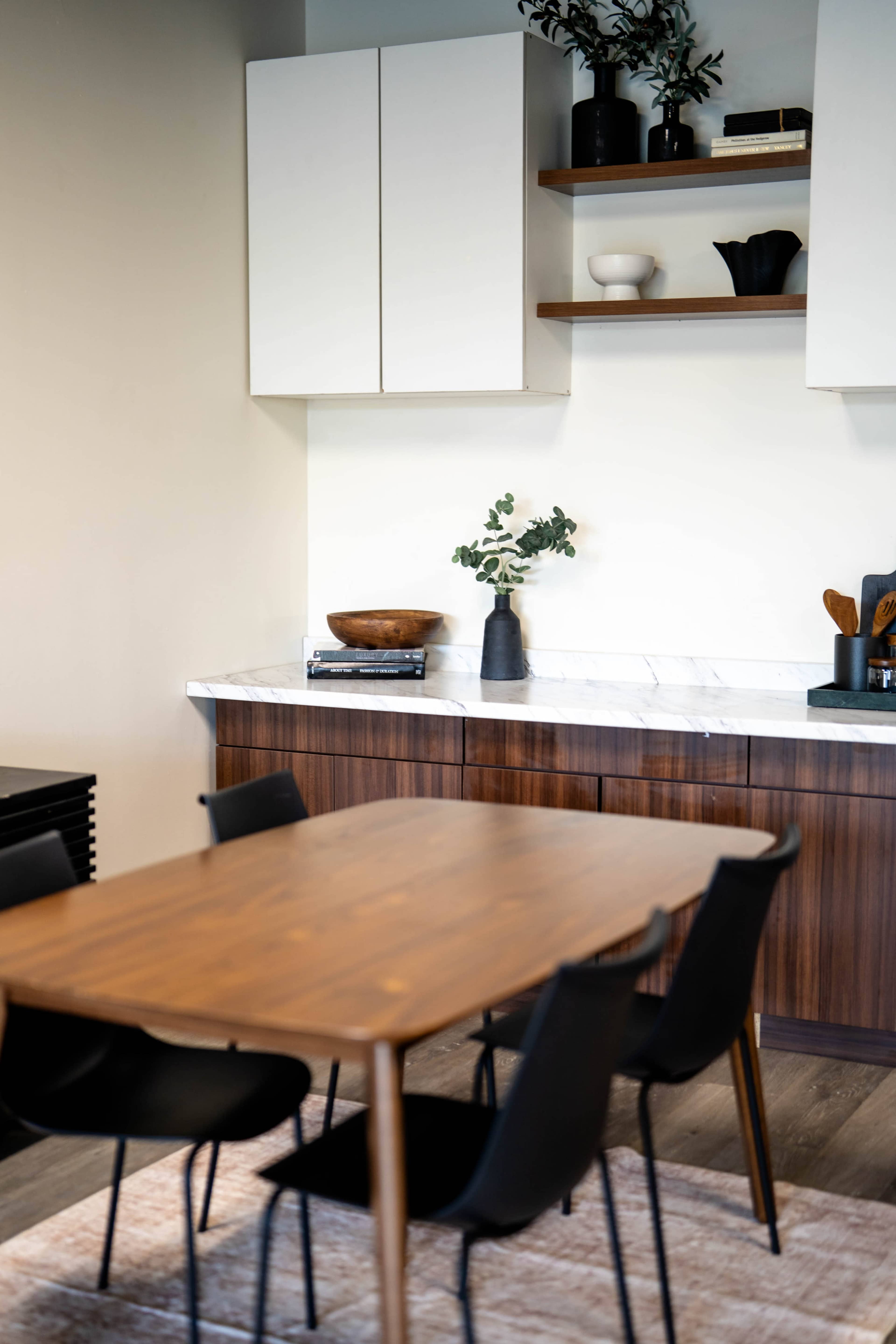 A modern dining area features a wooden table surrounded by black chairs, with a marble countertop and open shelves displaying decorative items.