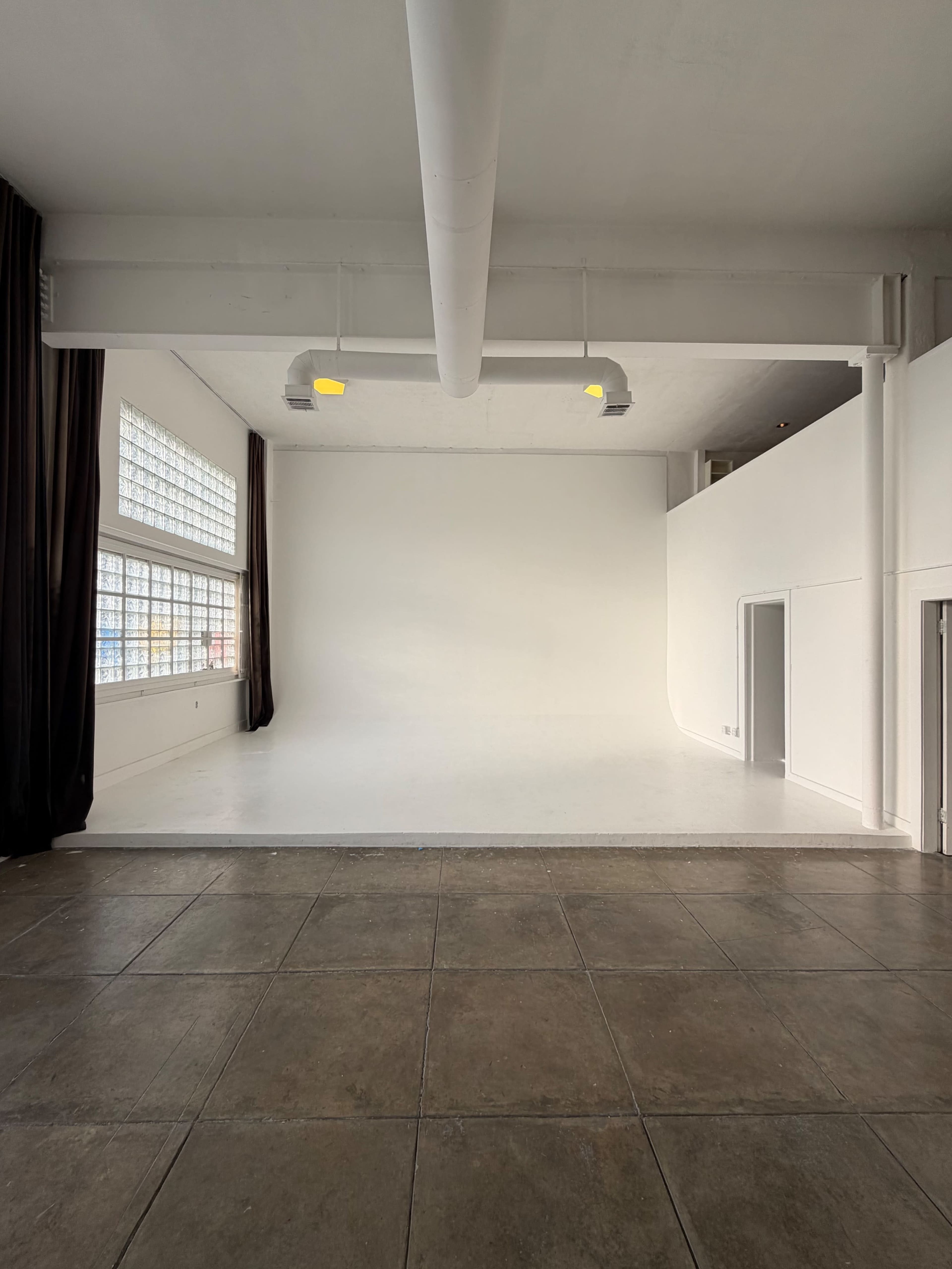 The image shows a spacious, empty studio with white walls, a light-colored floor, and large windows allowing natural light.