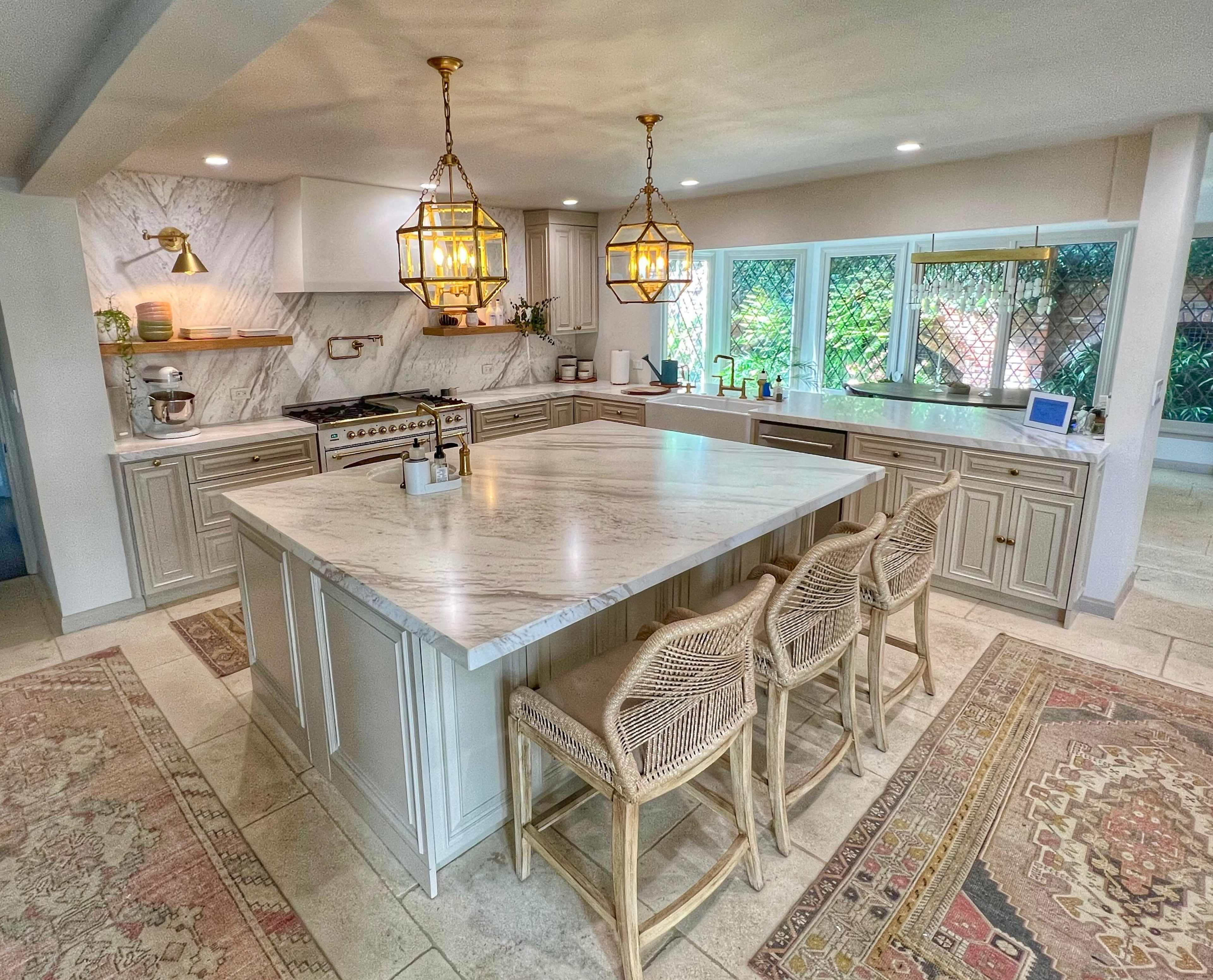 The kitchen features a large marble island with four rattan bar stools, stylish pendant lights, and modern appliances among light cabinetry and natural light coming from surrounding windows.