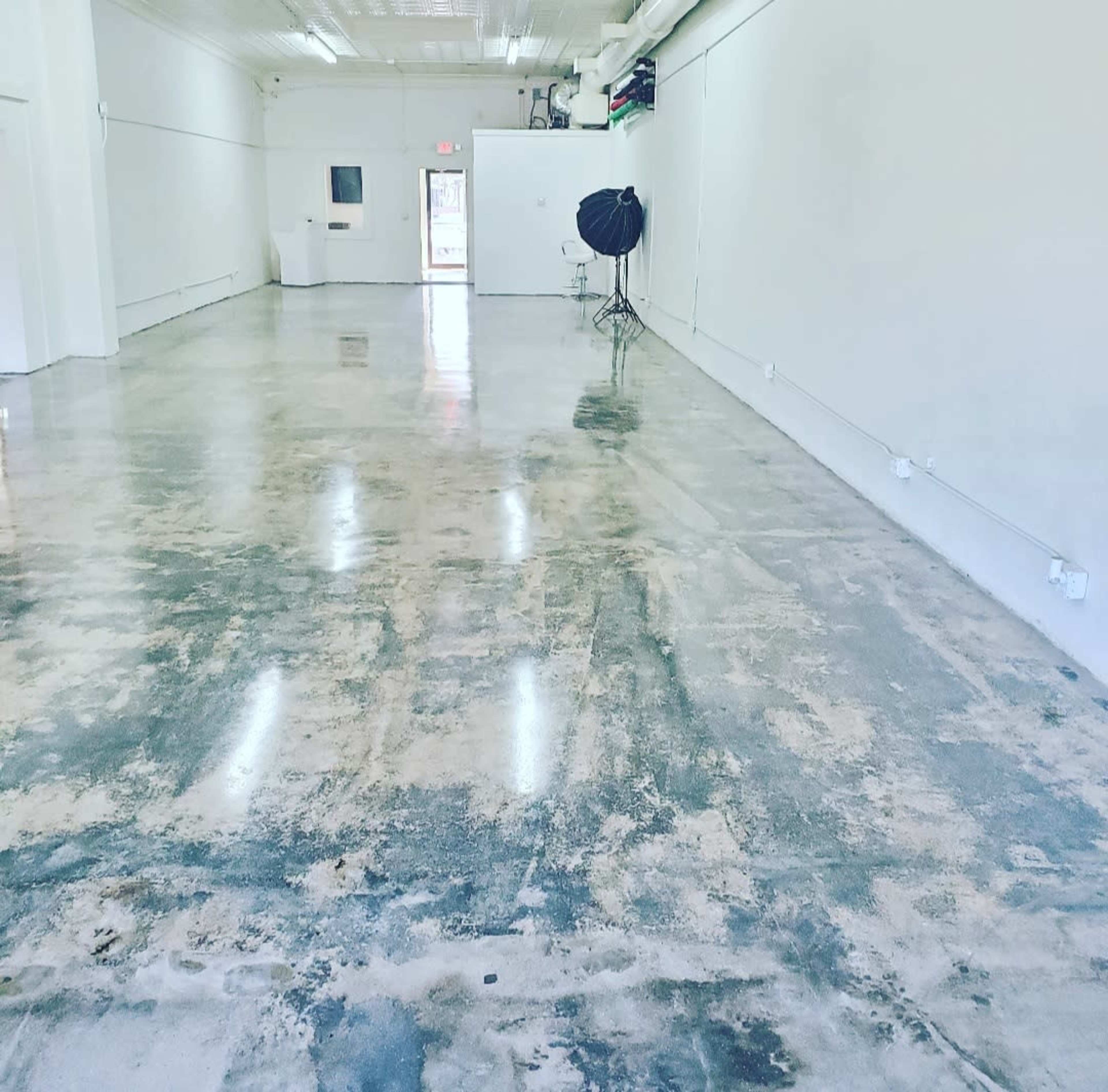The image shows a spacious, empty room with polished concrete flooring and white walls, illuminated by natural light.