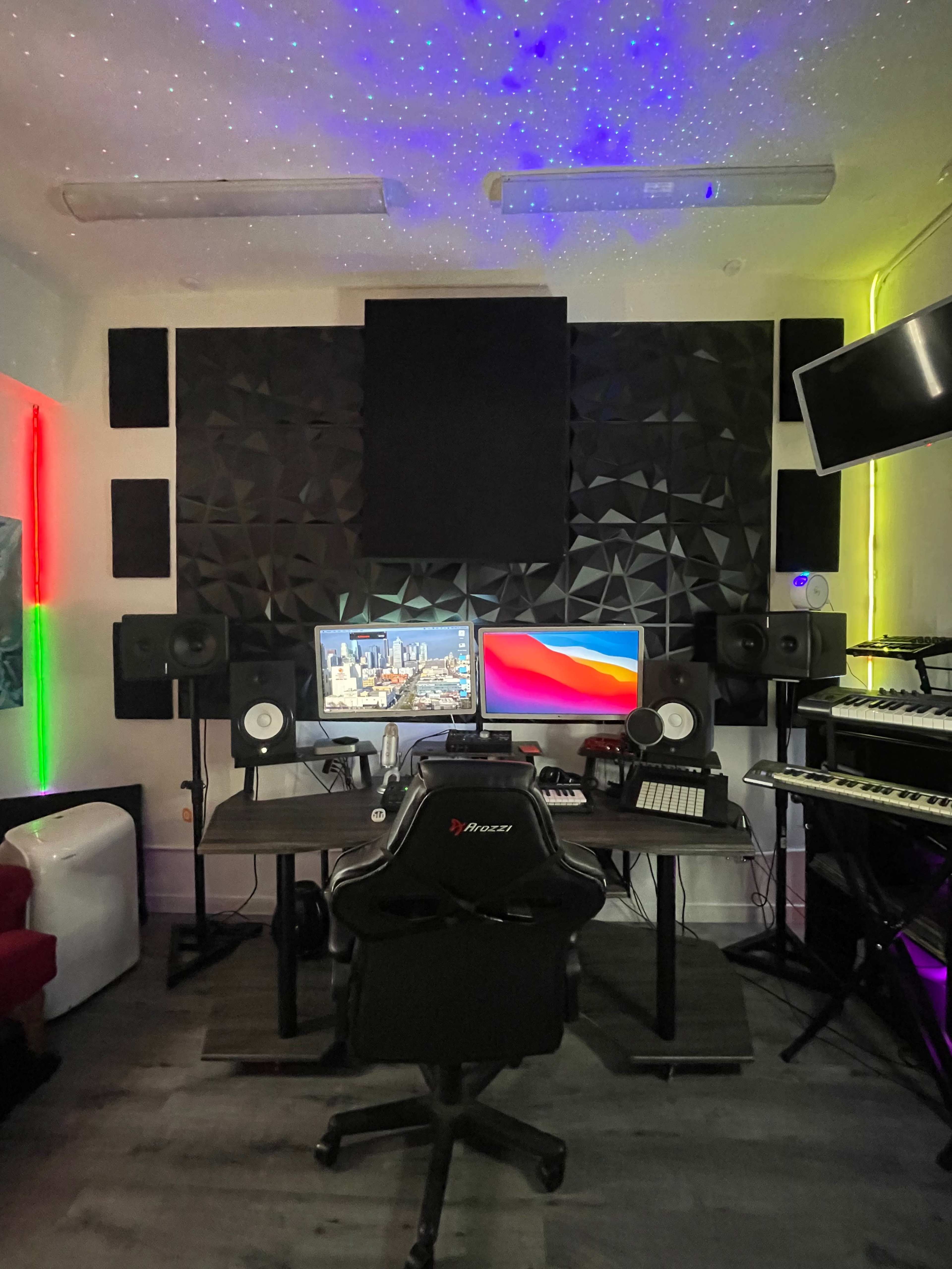 The image shows a music production studio featuring two computer monitors, surround speakers, a keyboard, and colorful LED lighting on the walls.