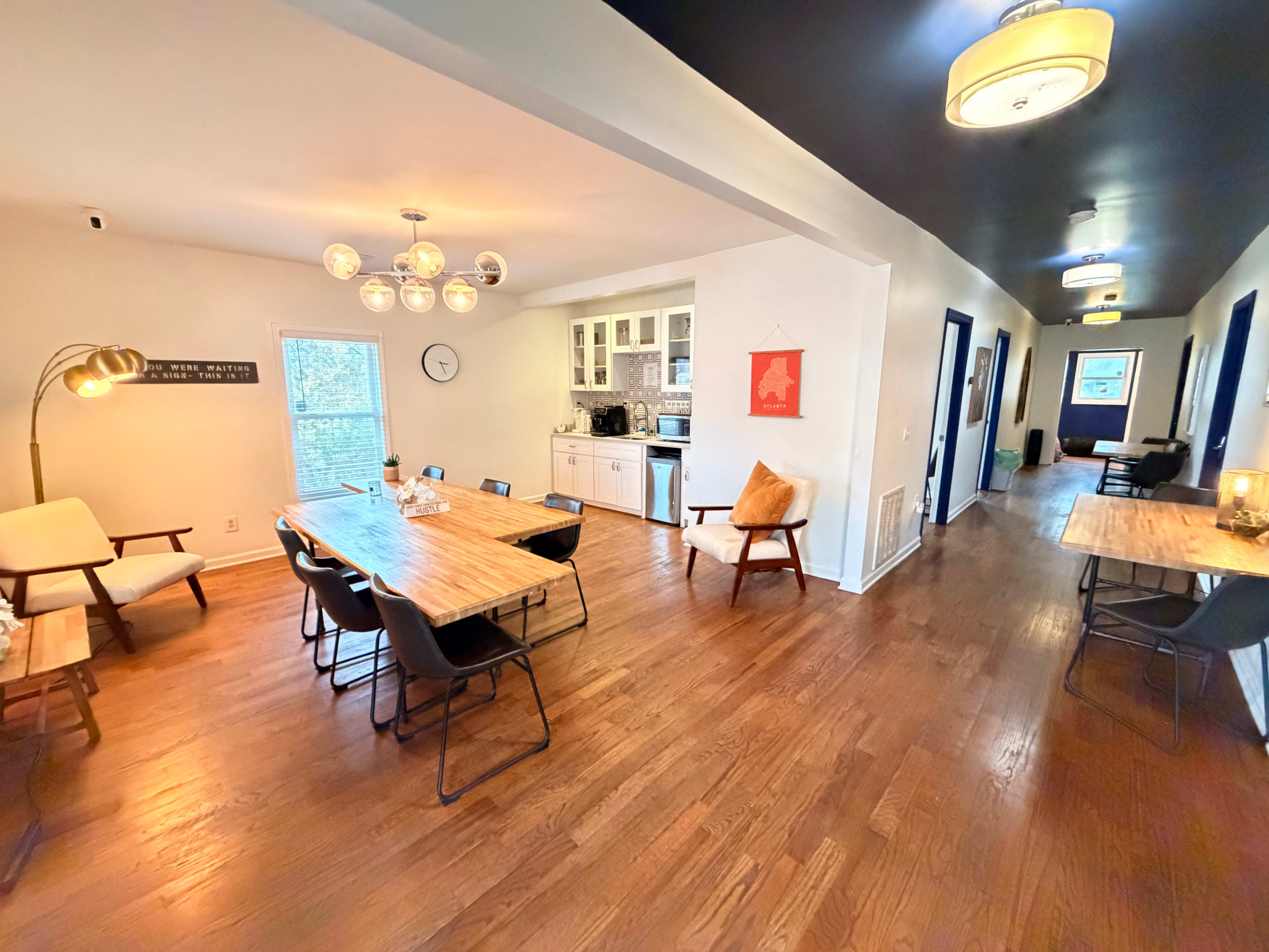 The image shows a spacious and modern dining area with wooden tables, chairs, and a view into a kitchen area, along with a hallway leading to another room.