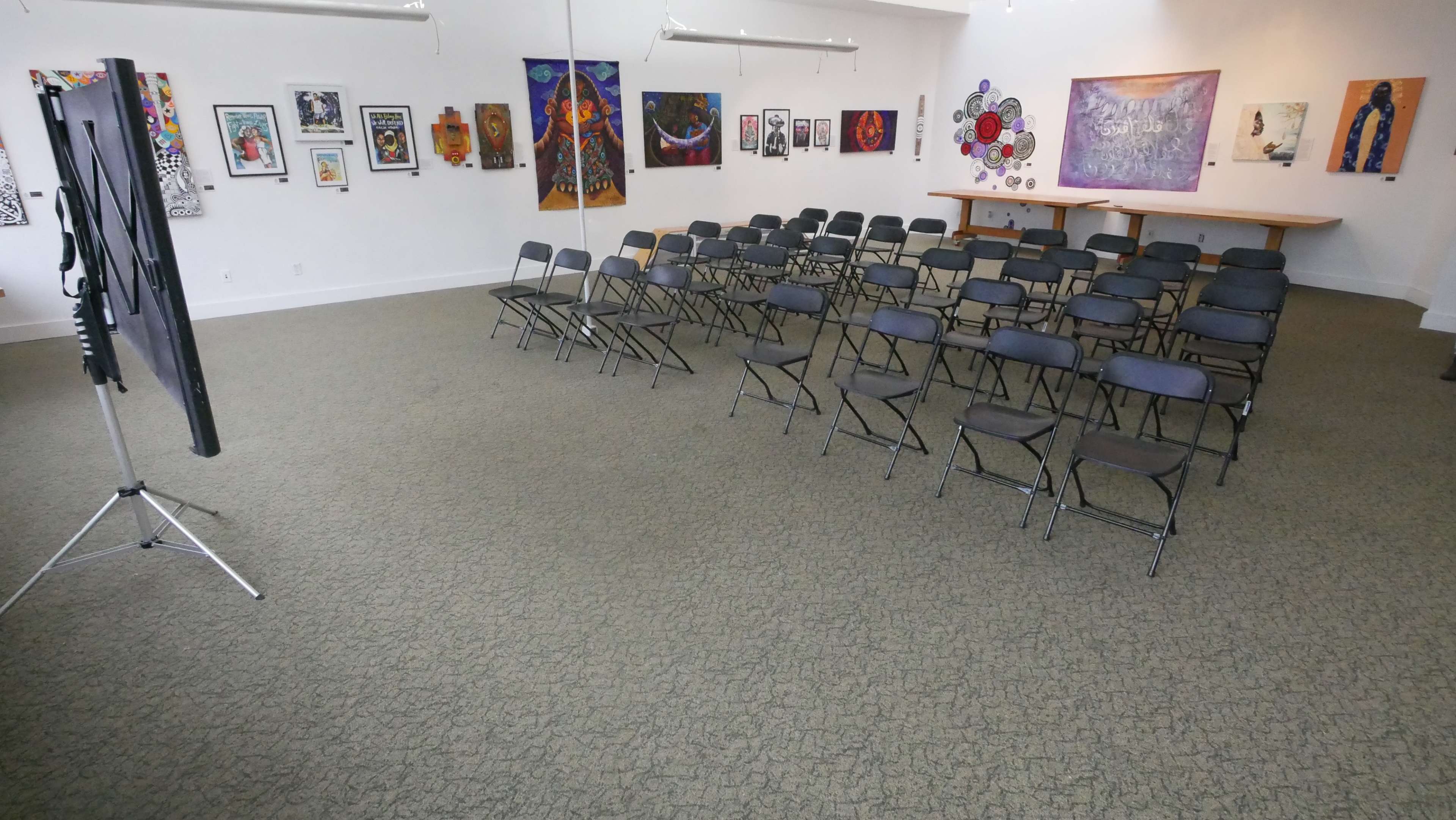 A spacious room with rows of black folding chairs facing a presentation area, adorned with various artworks on the walls.