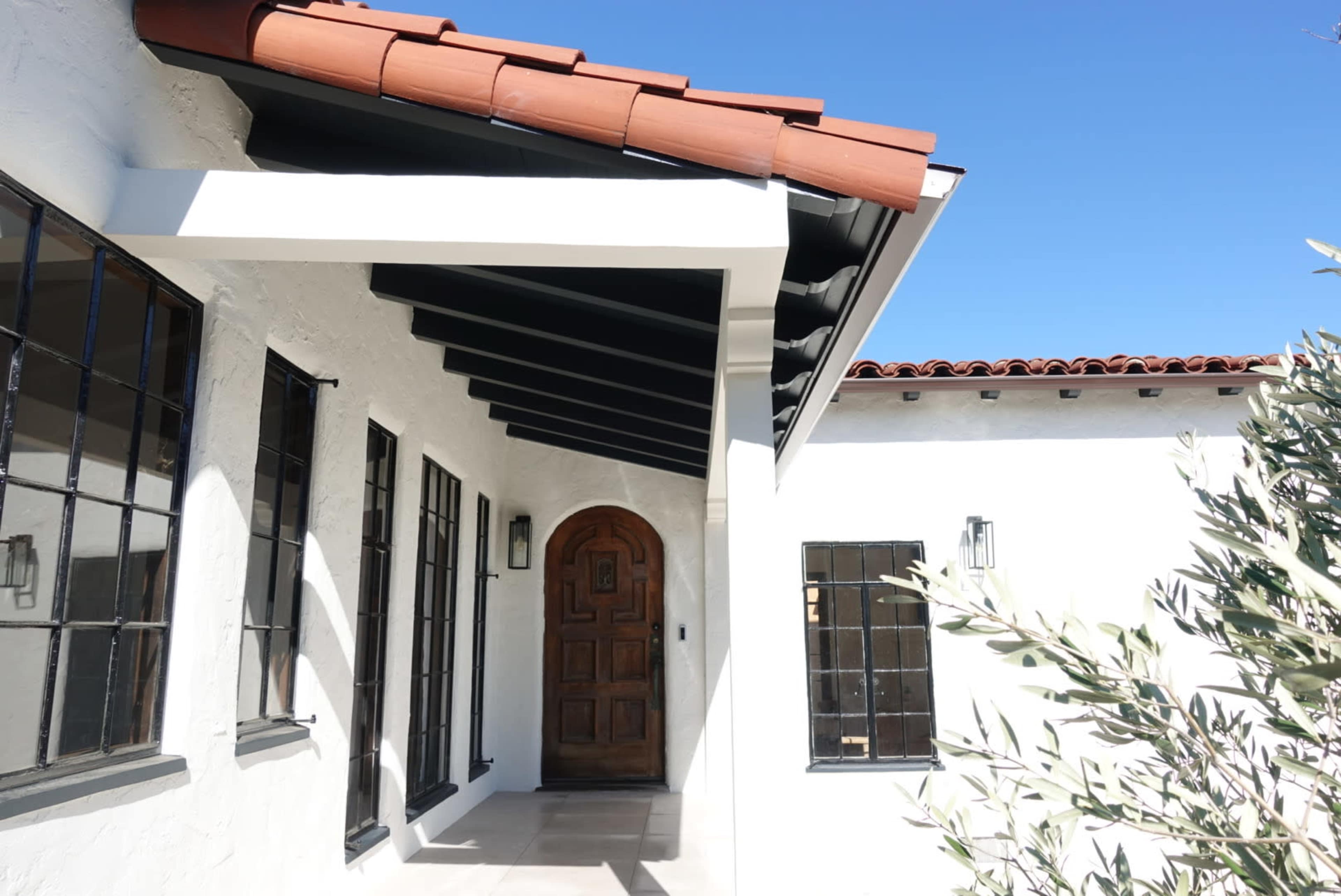 The image depicts a house entrance featuring a wooden door, black-framed windows, and a tiled roof under a clear blue sky.
