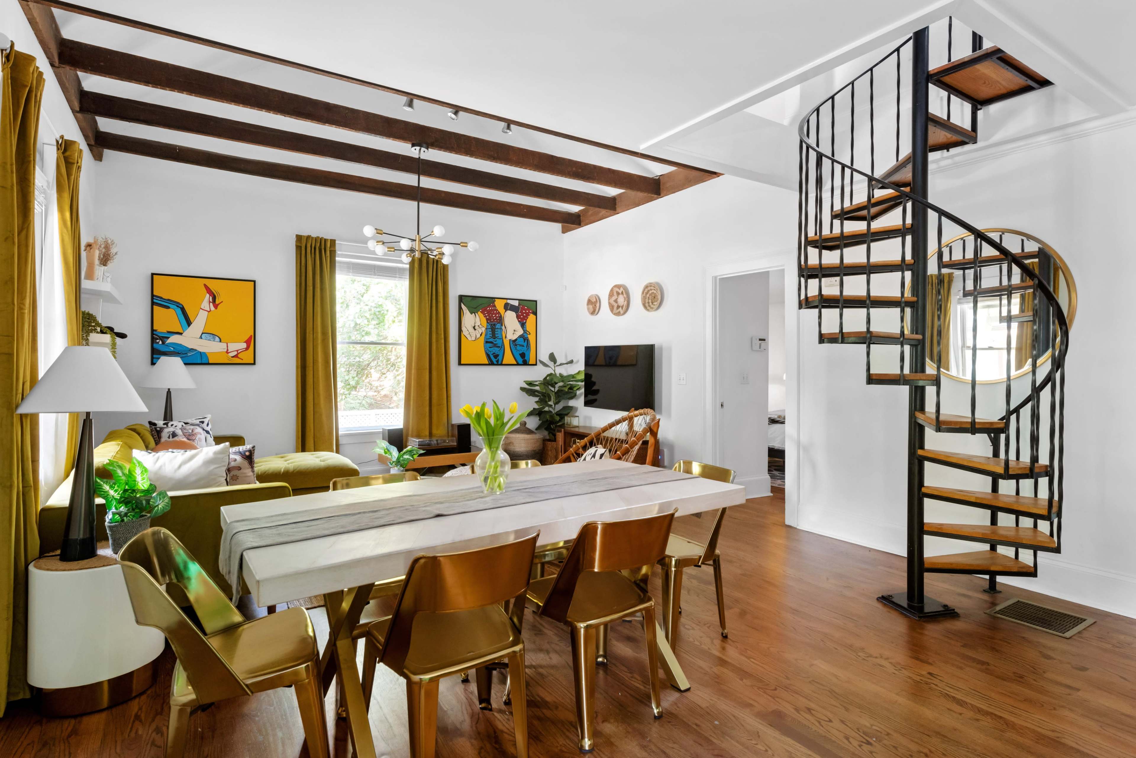 The image depicts a modern living space featuring a dining area with a long table, a spiral staircase, and artistic wall decor.