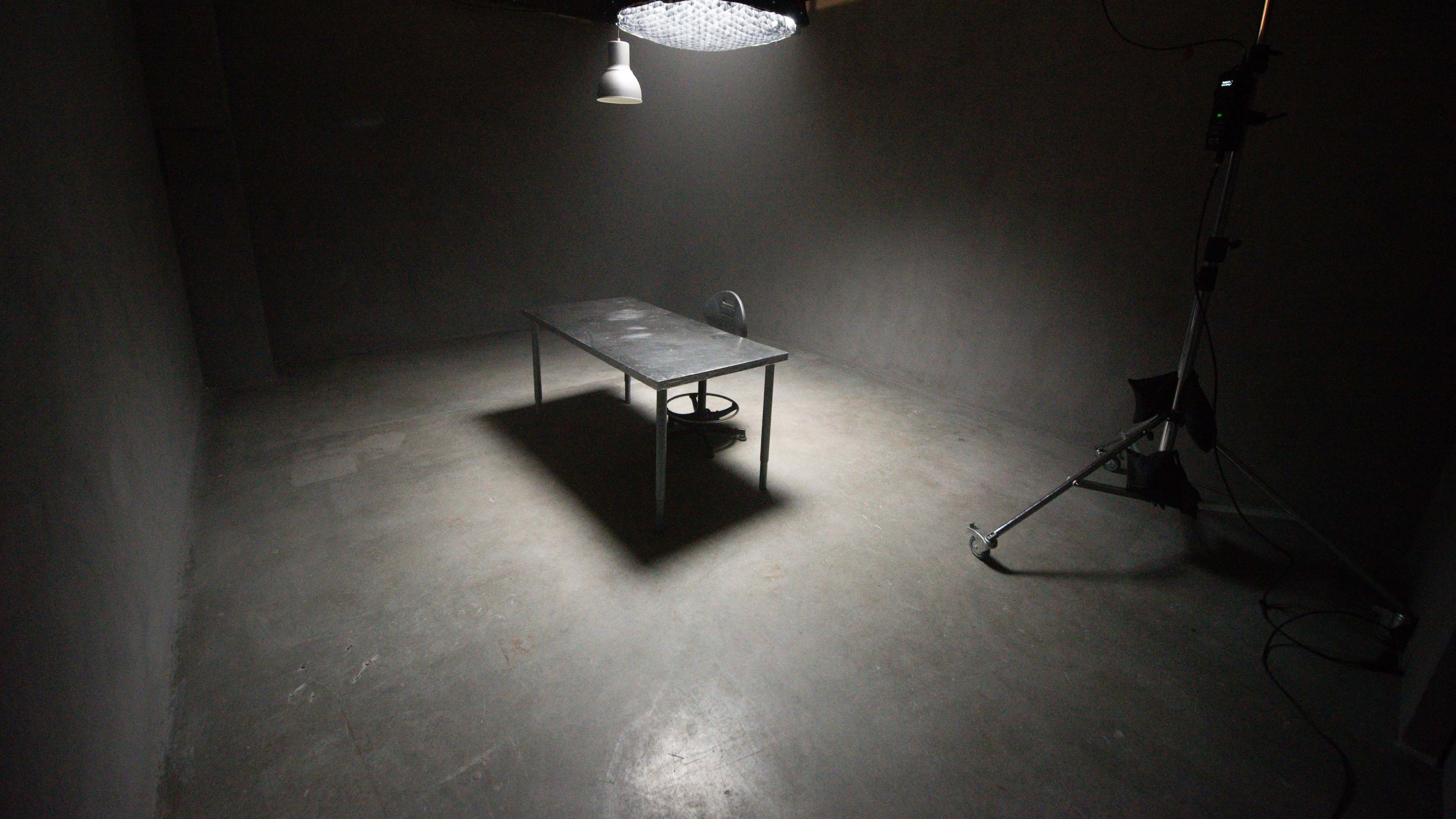 A sparse, dimly lit room contains a table and a fan under an overhead light.