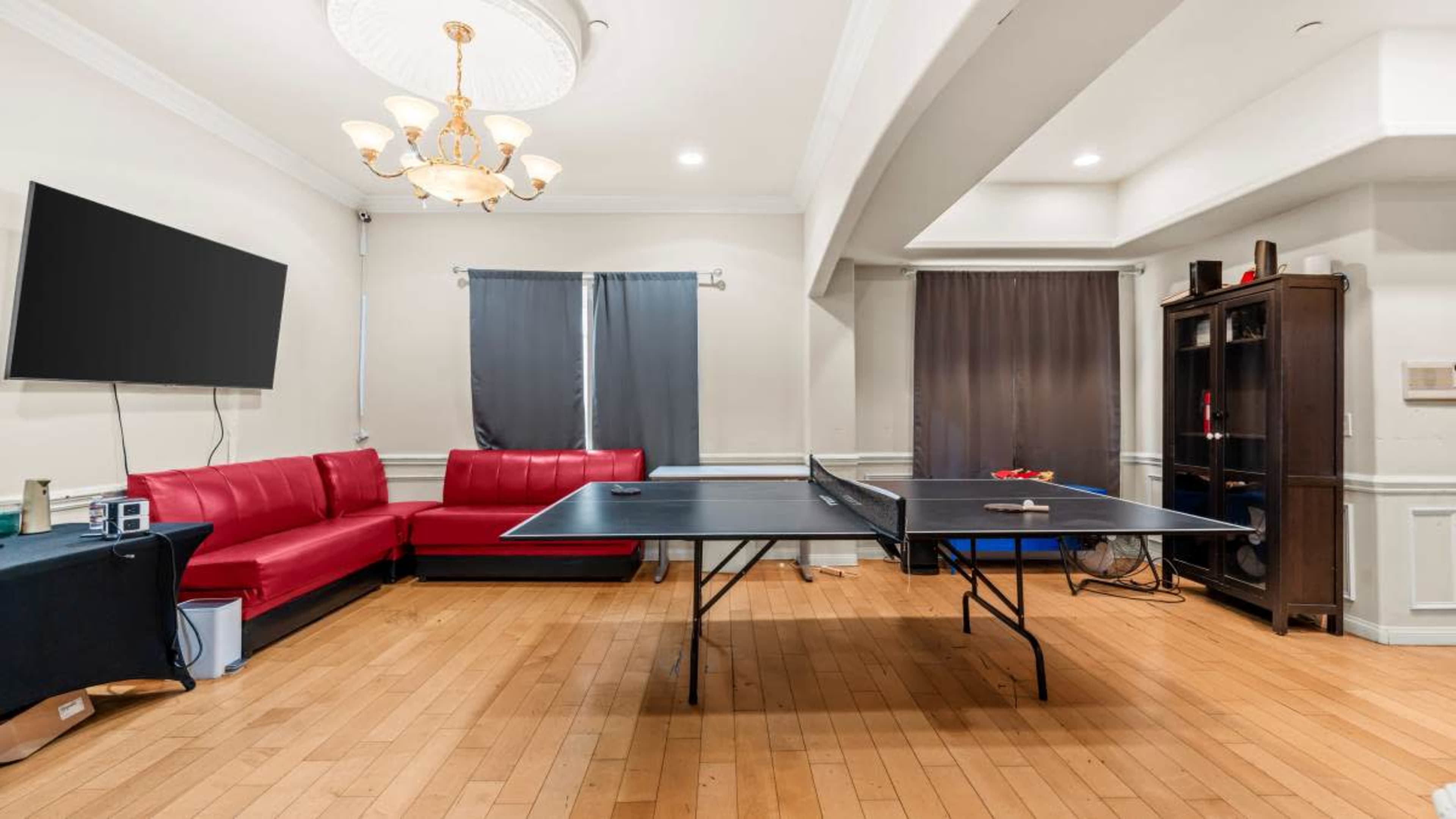 A ping pong table is set up in a room featuring a red leather sofa, a television on the wall, and gray curtains covering the windows.