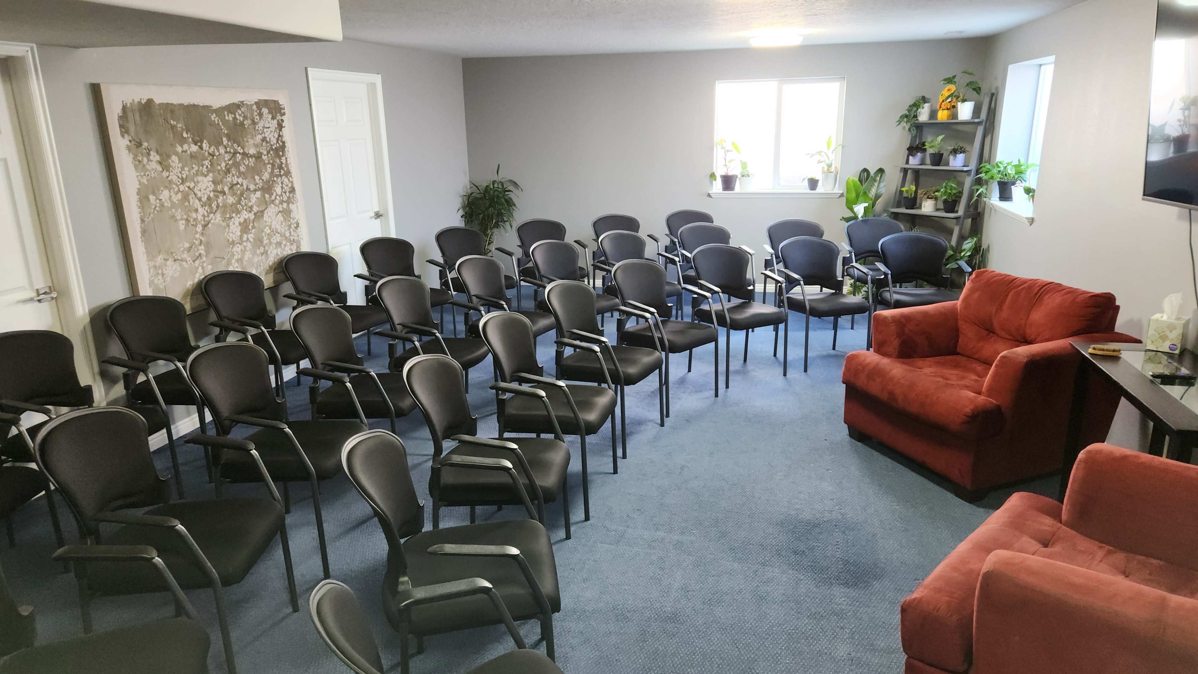 The room features multiple rows of black chairs arranged for seating, with a red armchair and a few potted plants visible in the corners.
