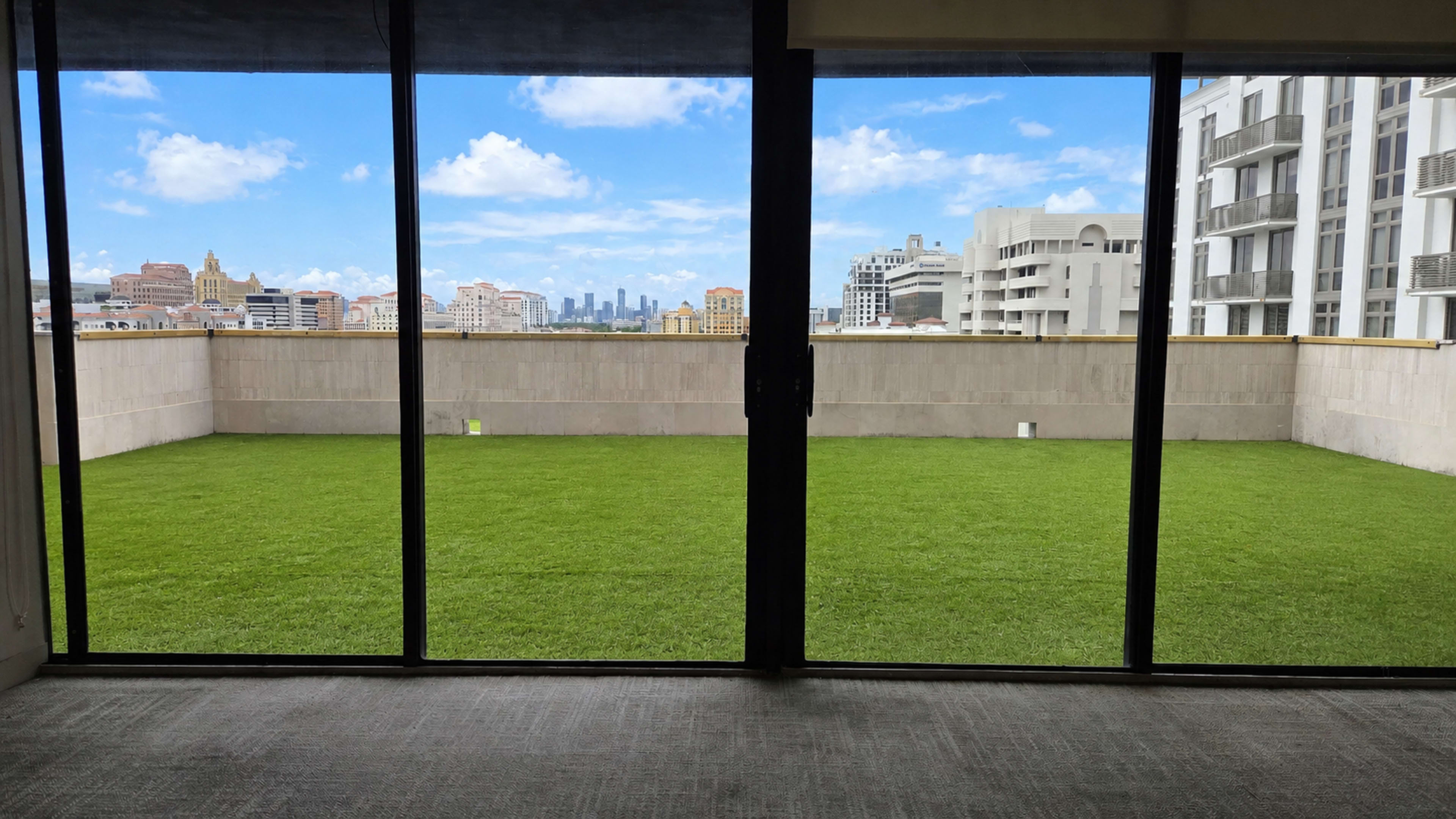 The image shows a glass door opening to a balcony with artificial grass, overlooking a city skyline under a blue sky with scattered clouds.