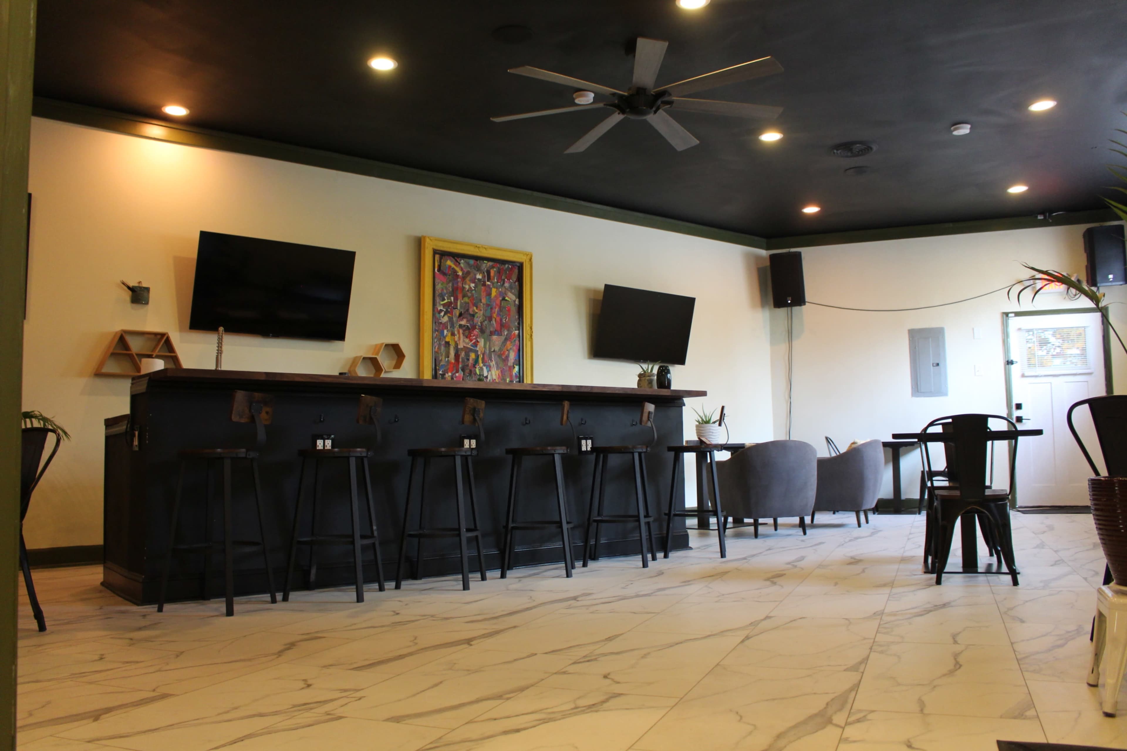 A modern bar area with a long black countertop, several bar stools, two wall-mounted televisions, and a seating area in a room with tiled flooring and dark ceiling.