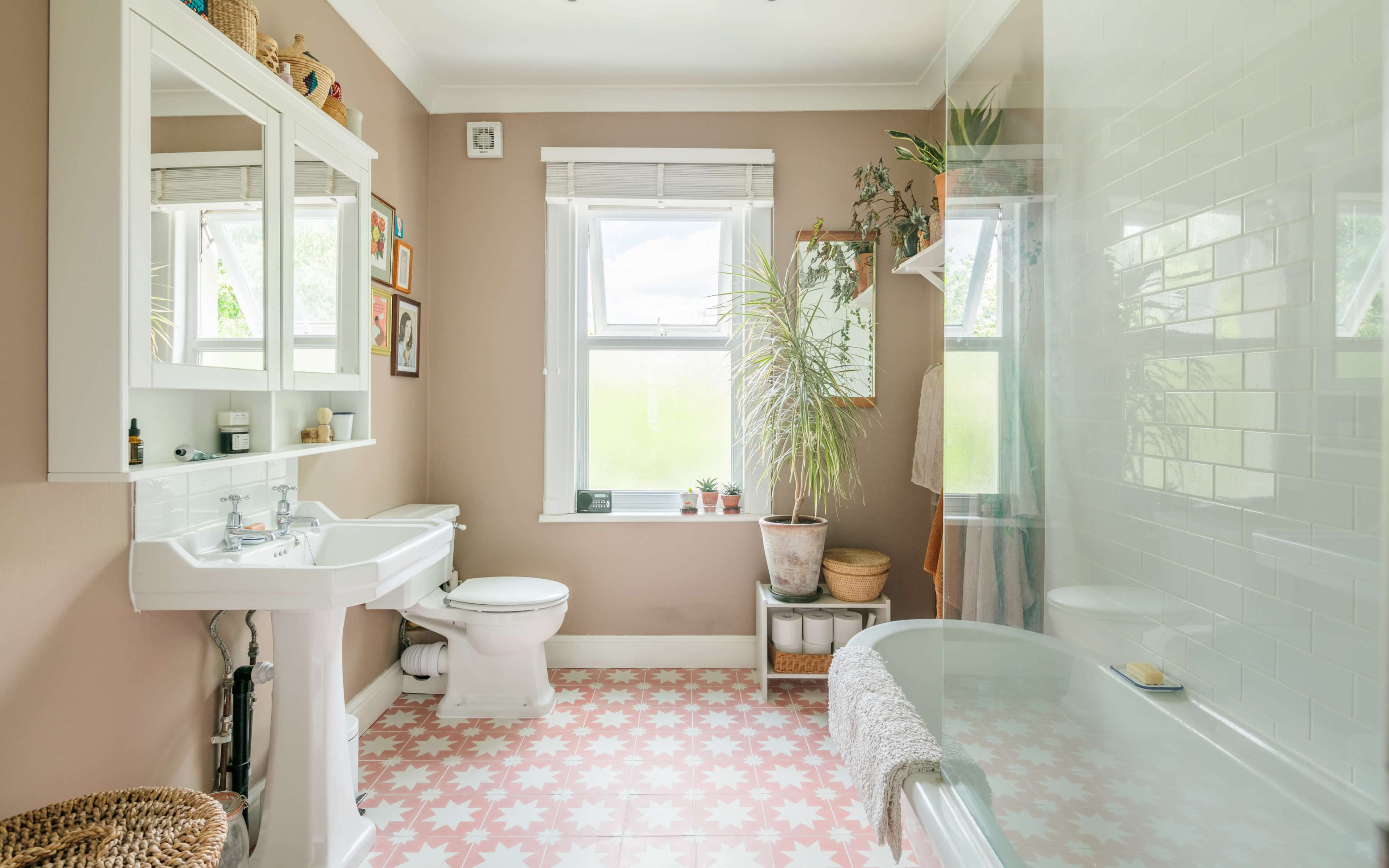A well-lit bathroom featuring a white sink and toilet, a bathtub, a large window with plants on the sill, and patterned pink tile flooring.