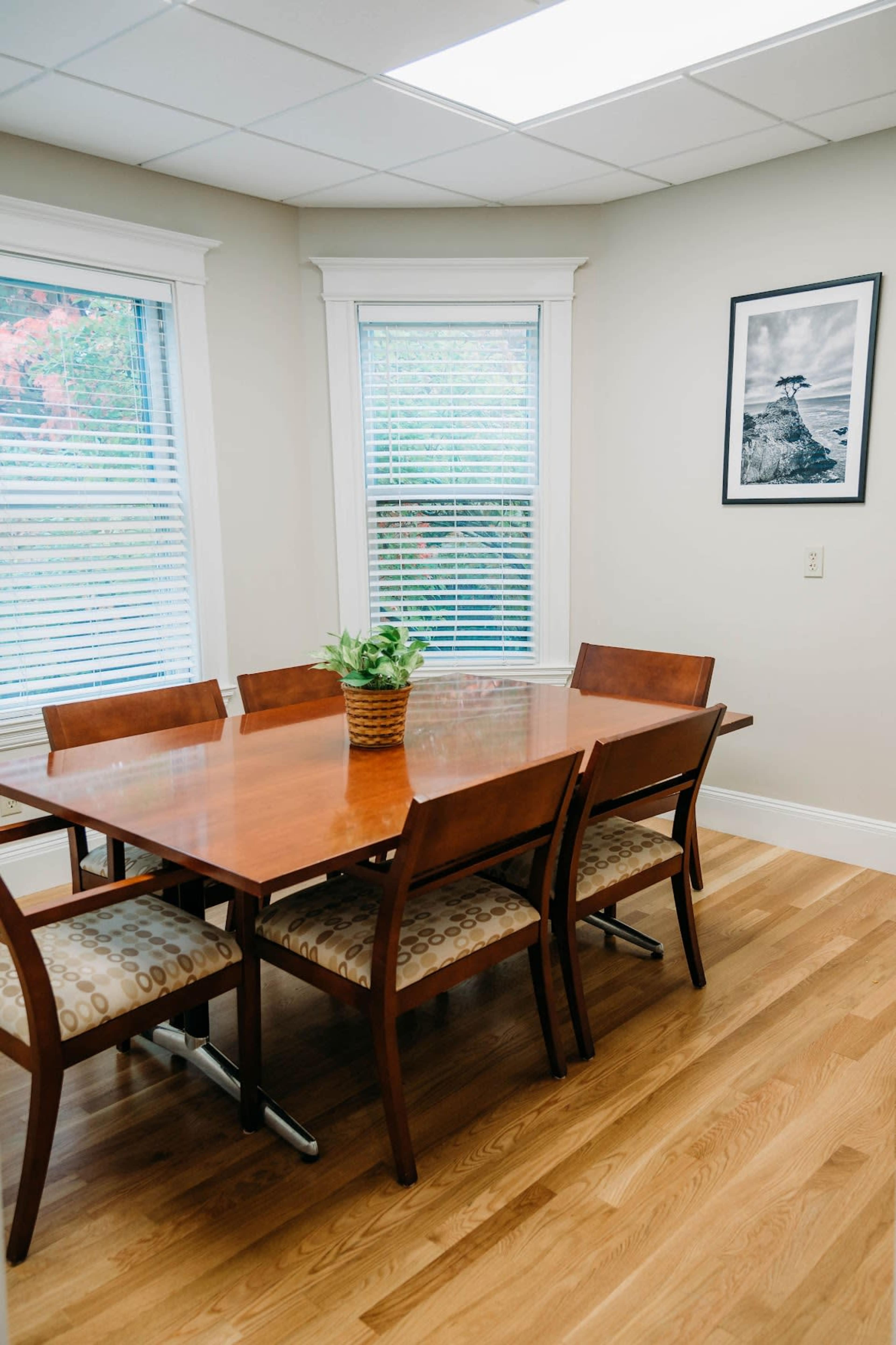 A wooden dining table with six chairs is positioned in a well-lit room featuring bay windows and a framed picture on the wall.