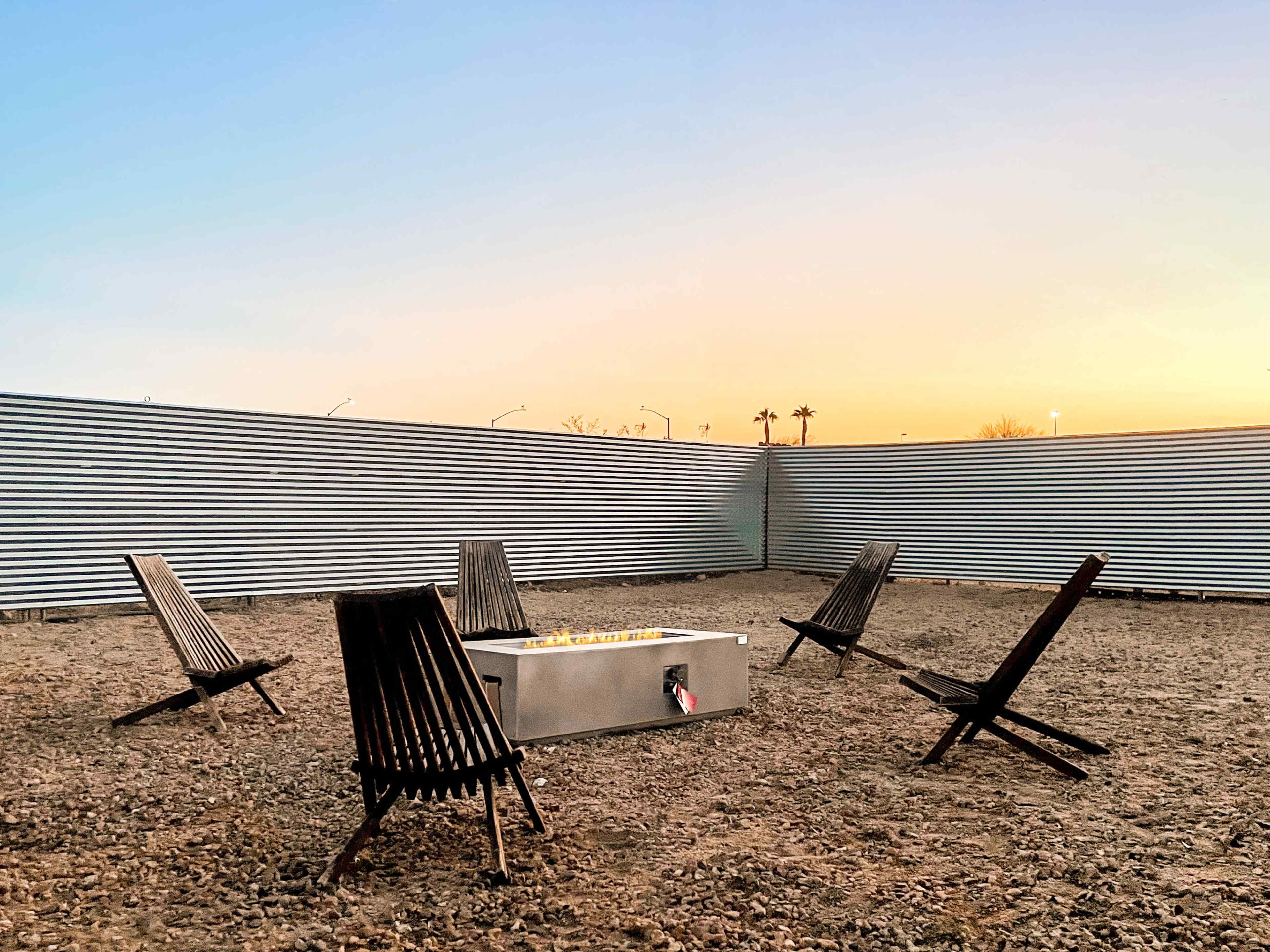 A circular arrangement of wooden chairs surrounds a metal fire pit in a fenced outdoor space at sunset.