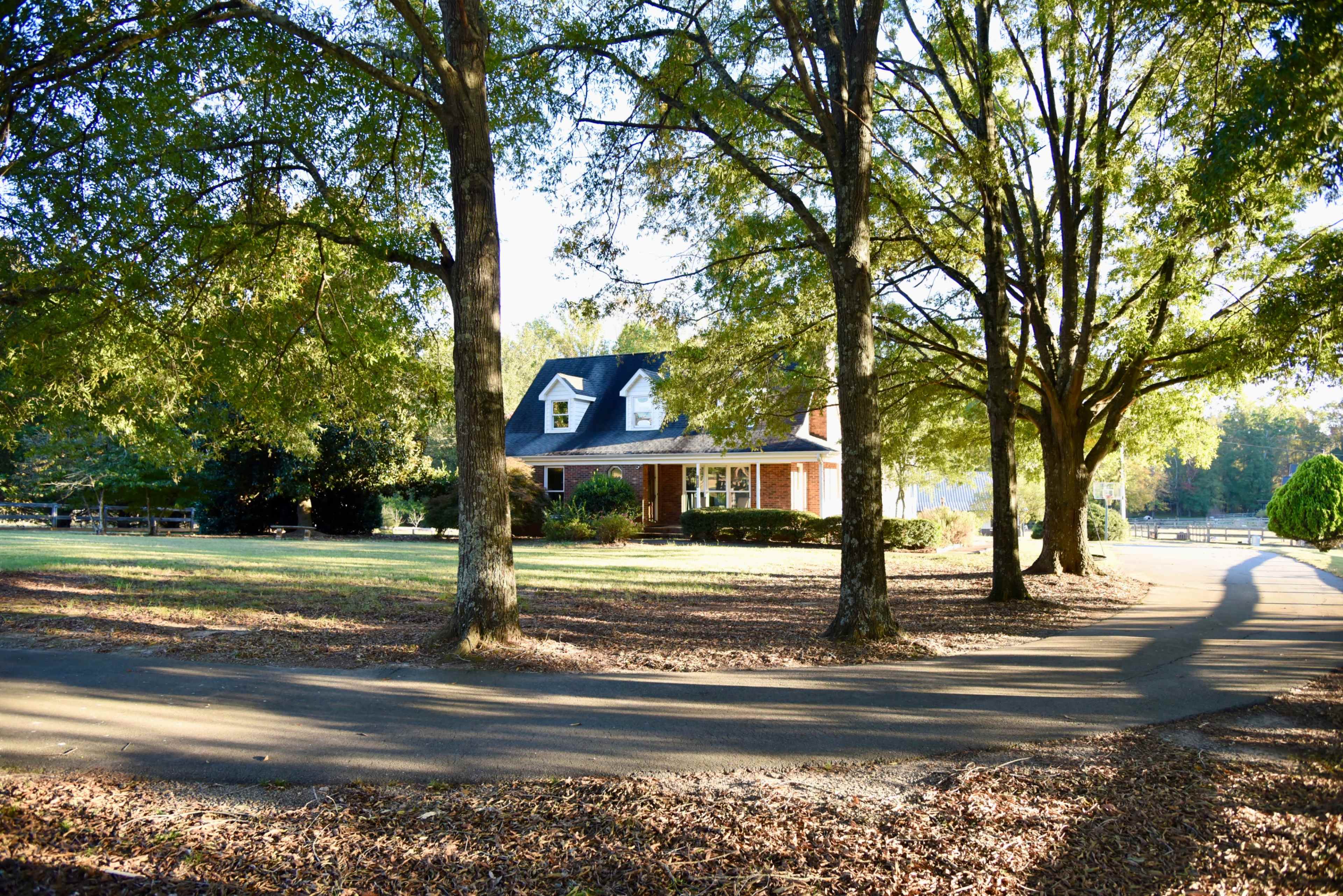 A brick house with a gabled roof is surrounded by large trees and a gravel driveway leading to a curved path.
