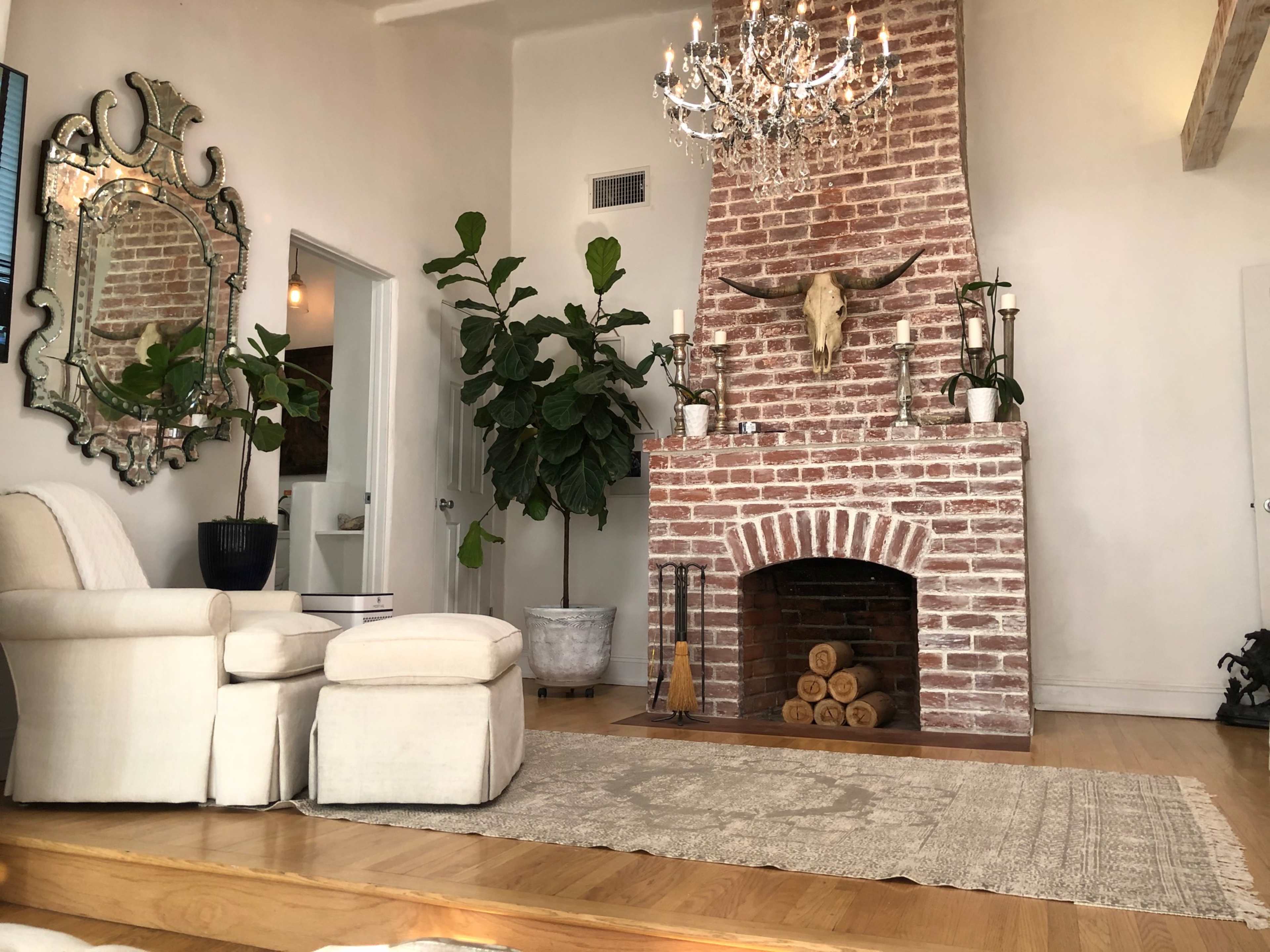 The image shows a living room with a brick fireplace, a chandelier, potted plants, and a comfortable seating area.