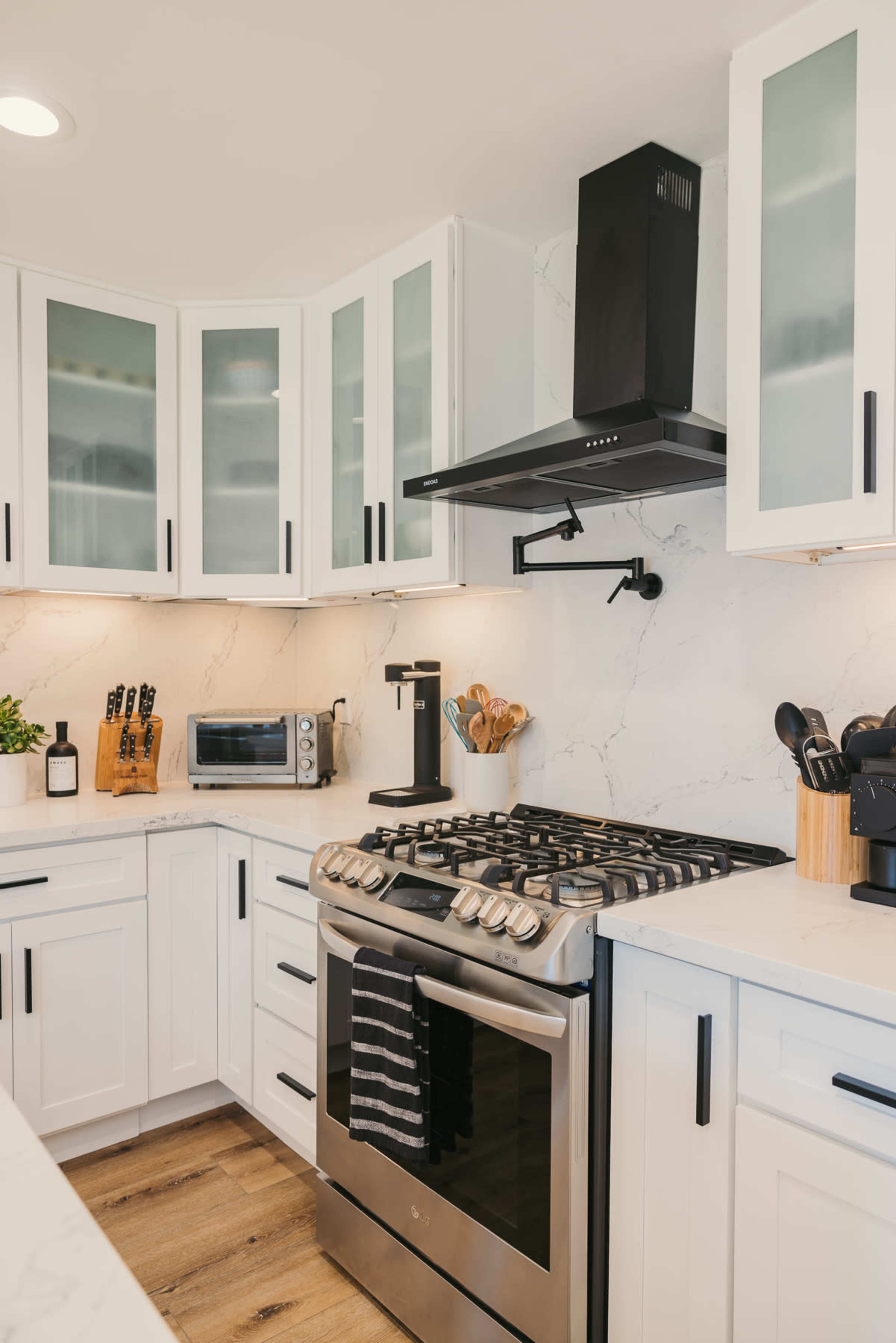 A modern kitchen features white cabinetry, a stainless steel gas range, and a range hood, with a countertop and backsplash made of marble.