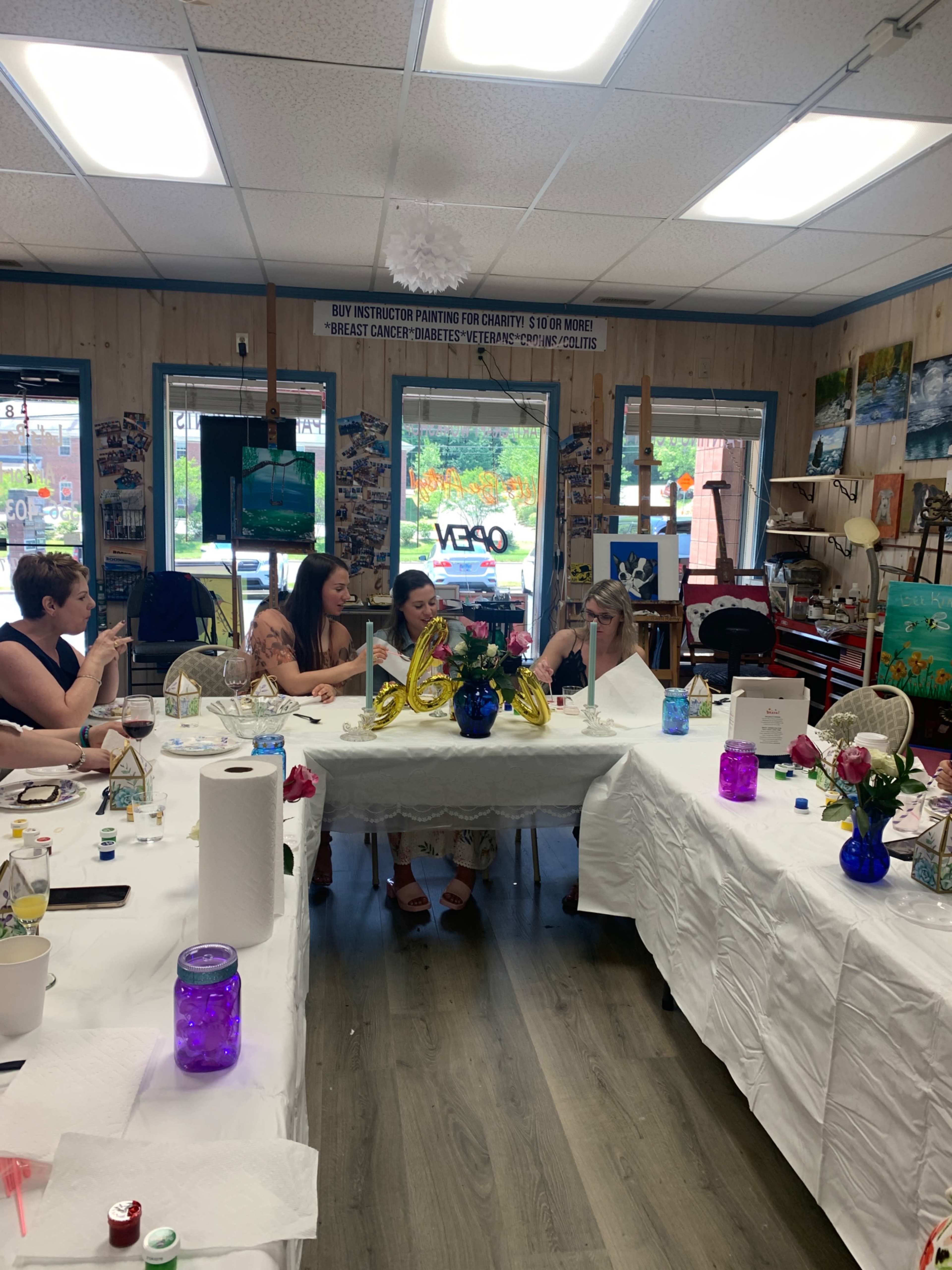 A group of women sitting at tables in an art studio, engaged in a painting activity while surrounded by various art supplies and decor.