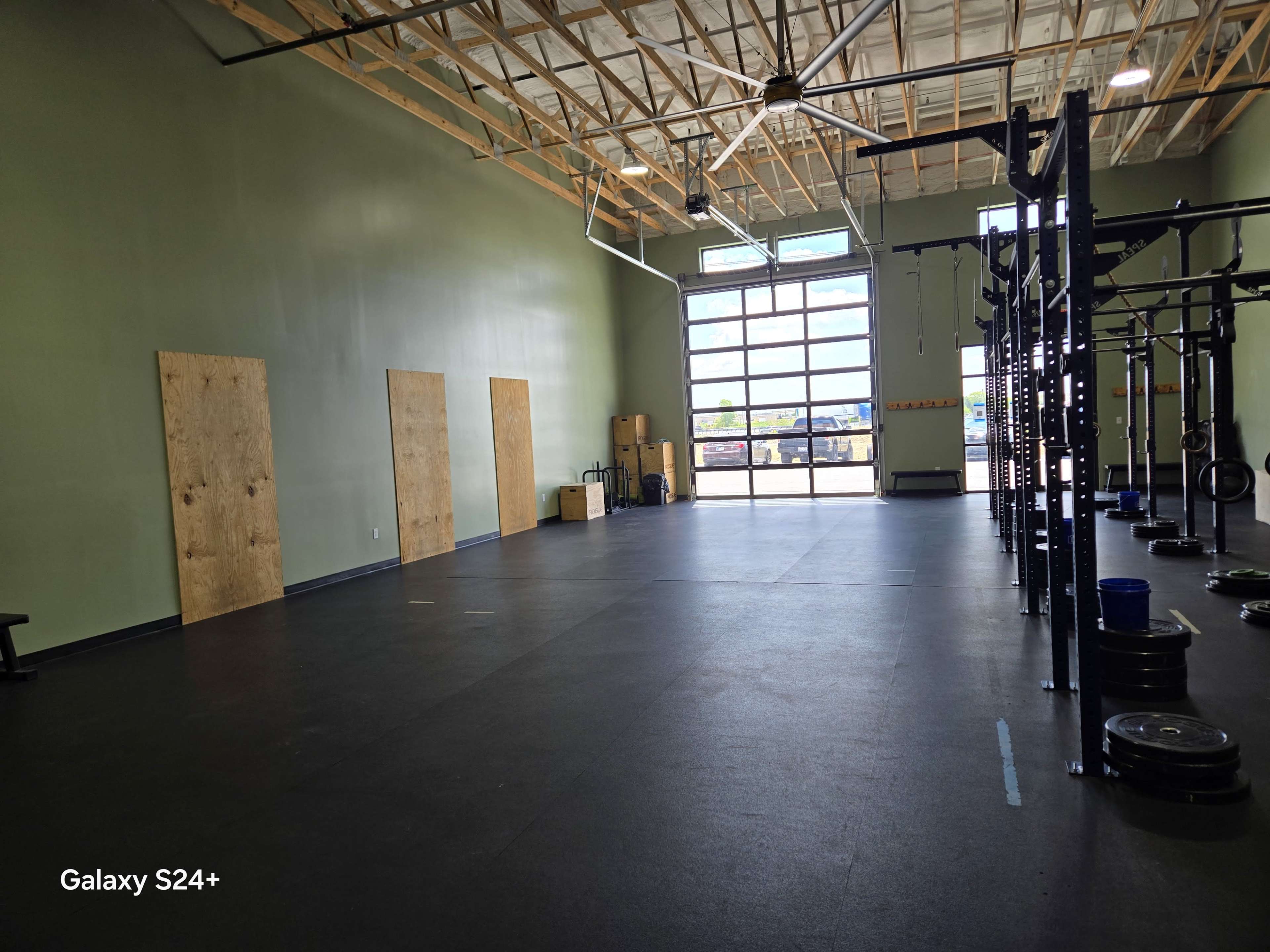 The image shows a large, empty gym space featuring a black rubber floor, a set of weightlifting equipment on one side, and wooden boards covering windows on the opposite wall.