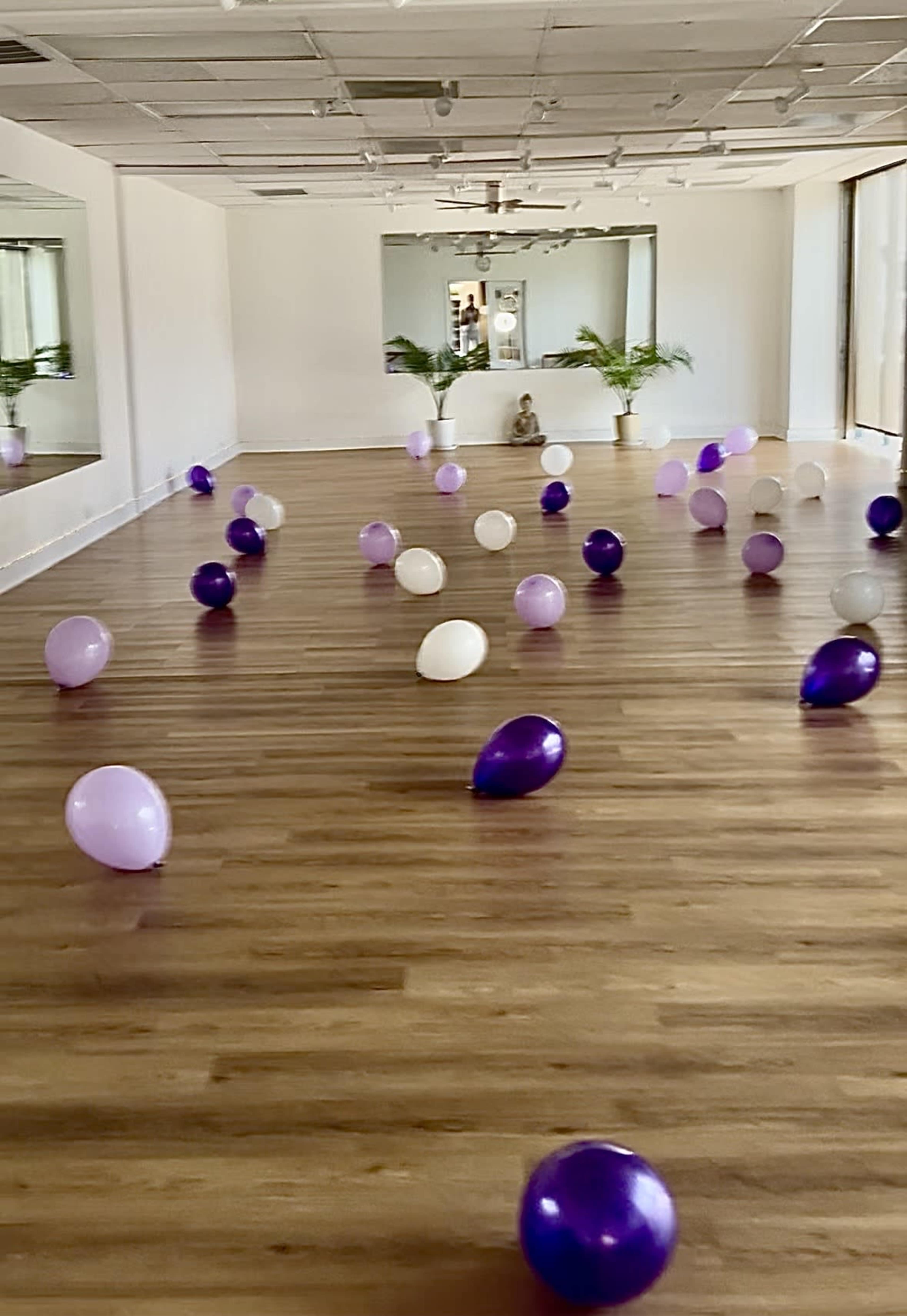 The image shows a spacious, empty room with wooden flooring scattered with purple, lavender, and white balloons.