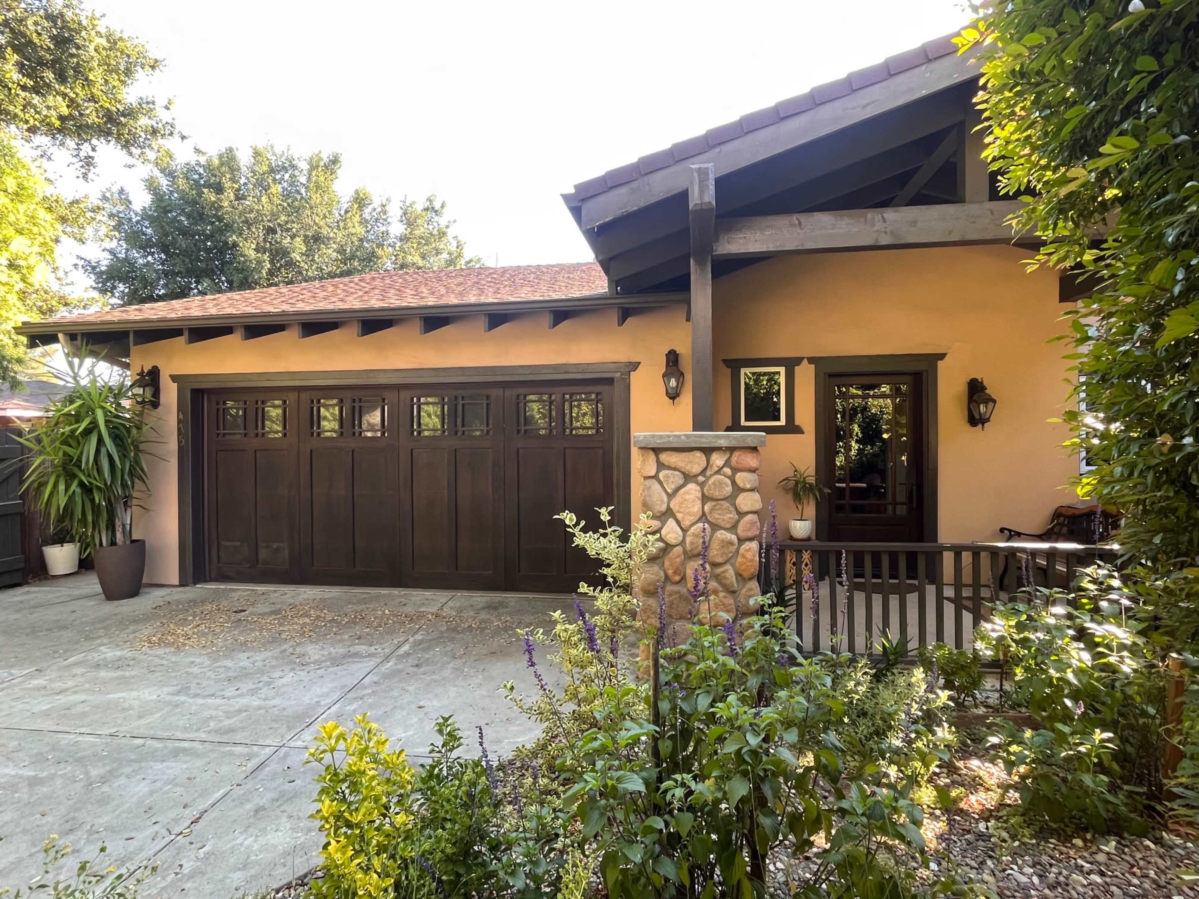 A suburban home features a stone and wooden garage door, with a landscaped front yard that includes various plants and shrubs.