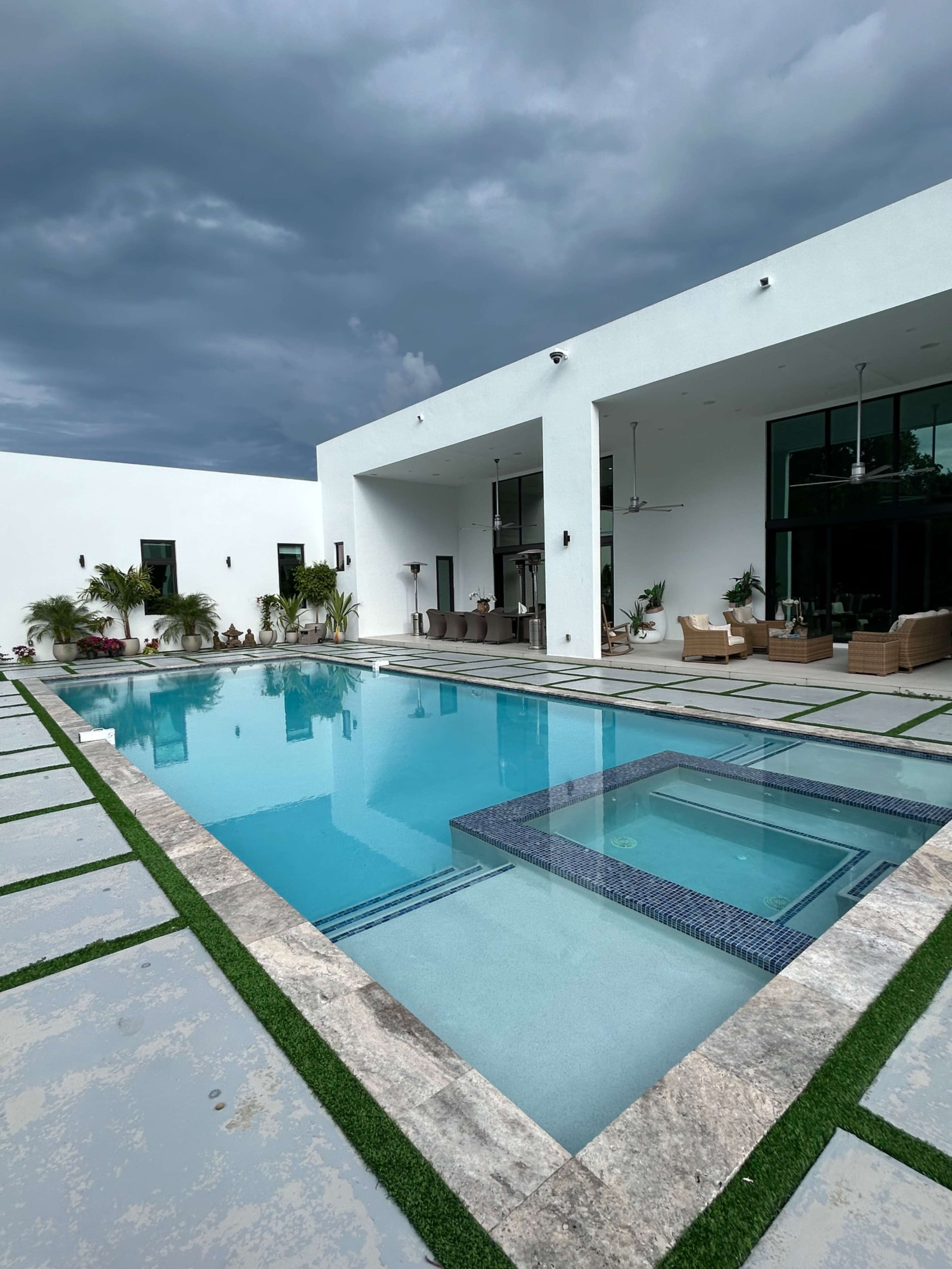 A modern house features a rectangular swimming pool surrounded by stone tiles and grass, with large windows and a covered seating area in the background.