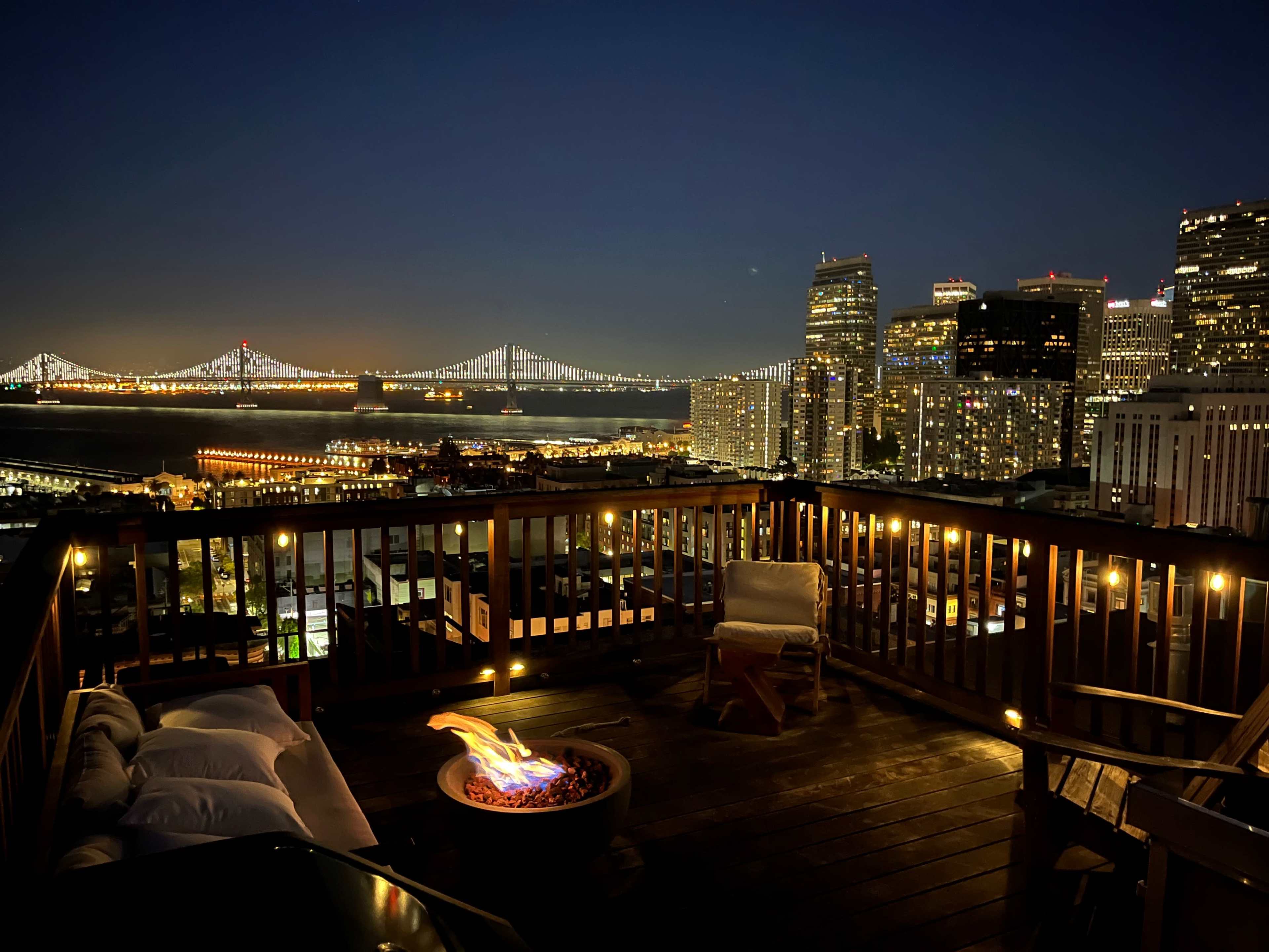 A rooftop terrace features a fire pit surrounded by seating, overlooking a city skyline illuminated at night.