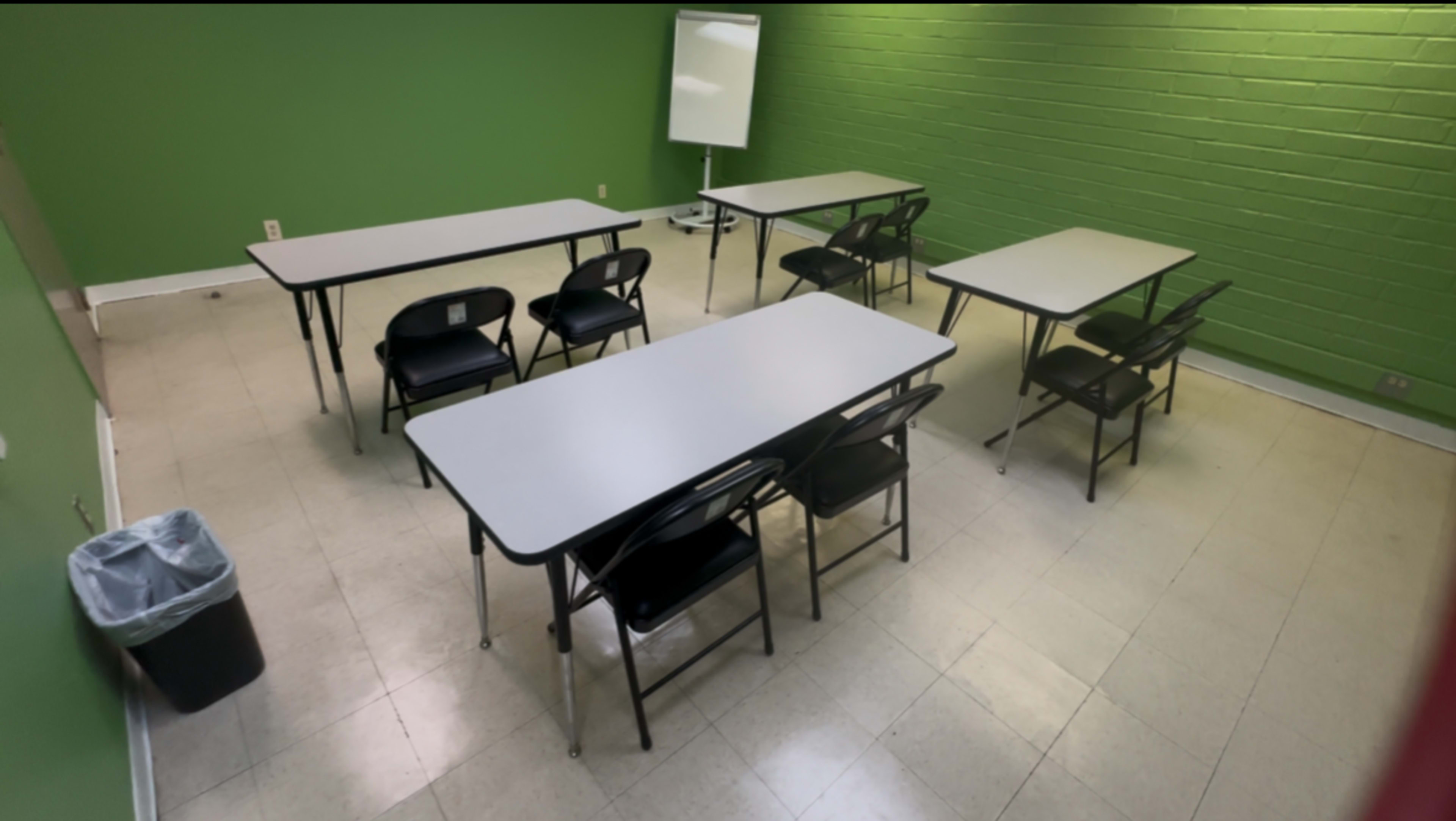 The image shows a small classroom with green walls, featuring six chairs arranged around two tables, along with a trash bin and a whiteboard in the corner.