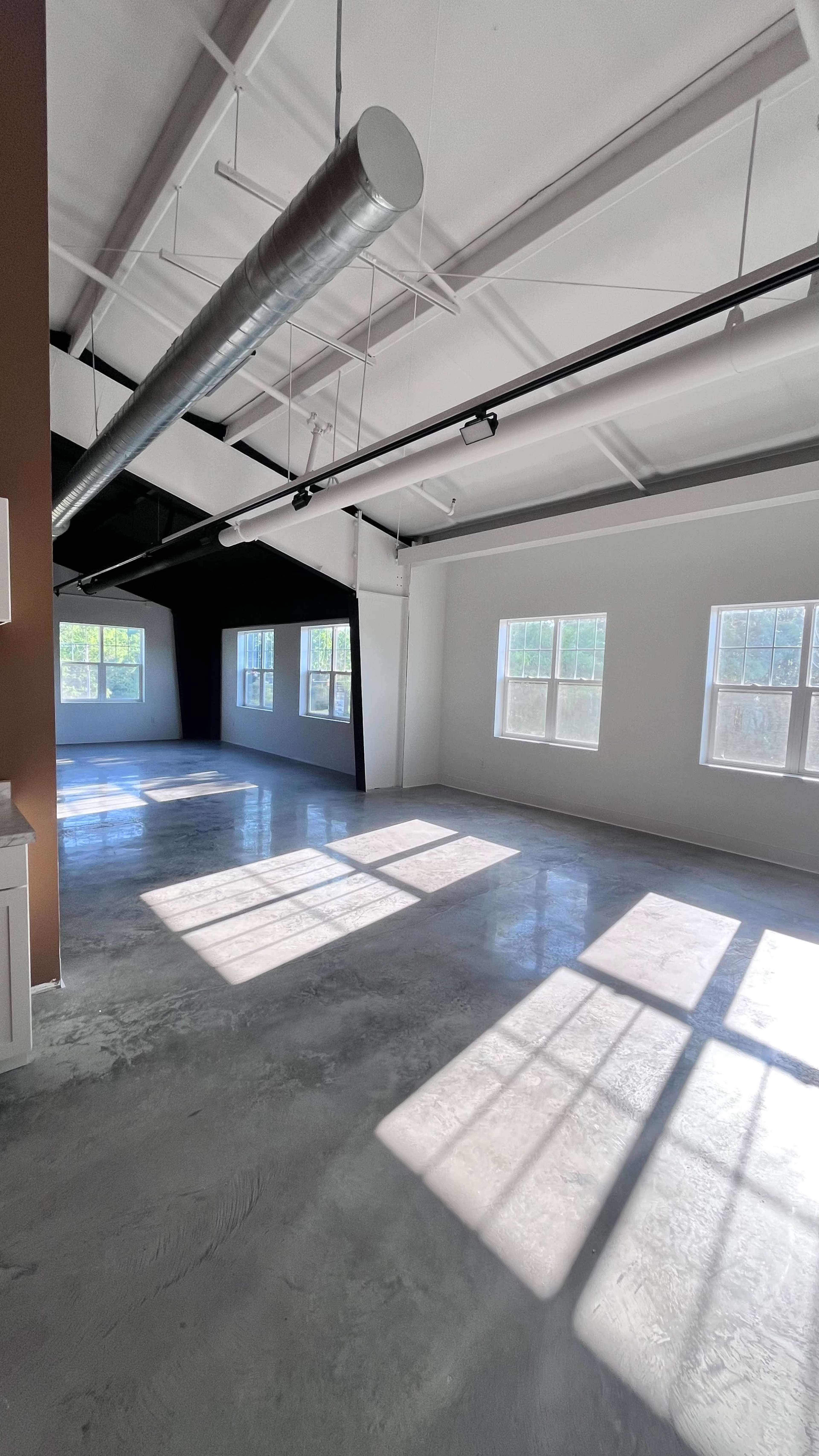 The image shows a large, empty room with concrete flooring and several windows letting in natural light.