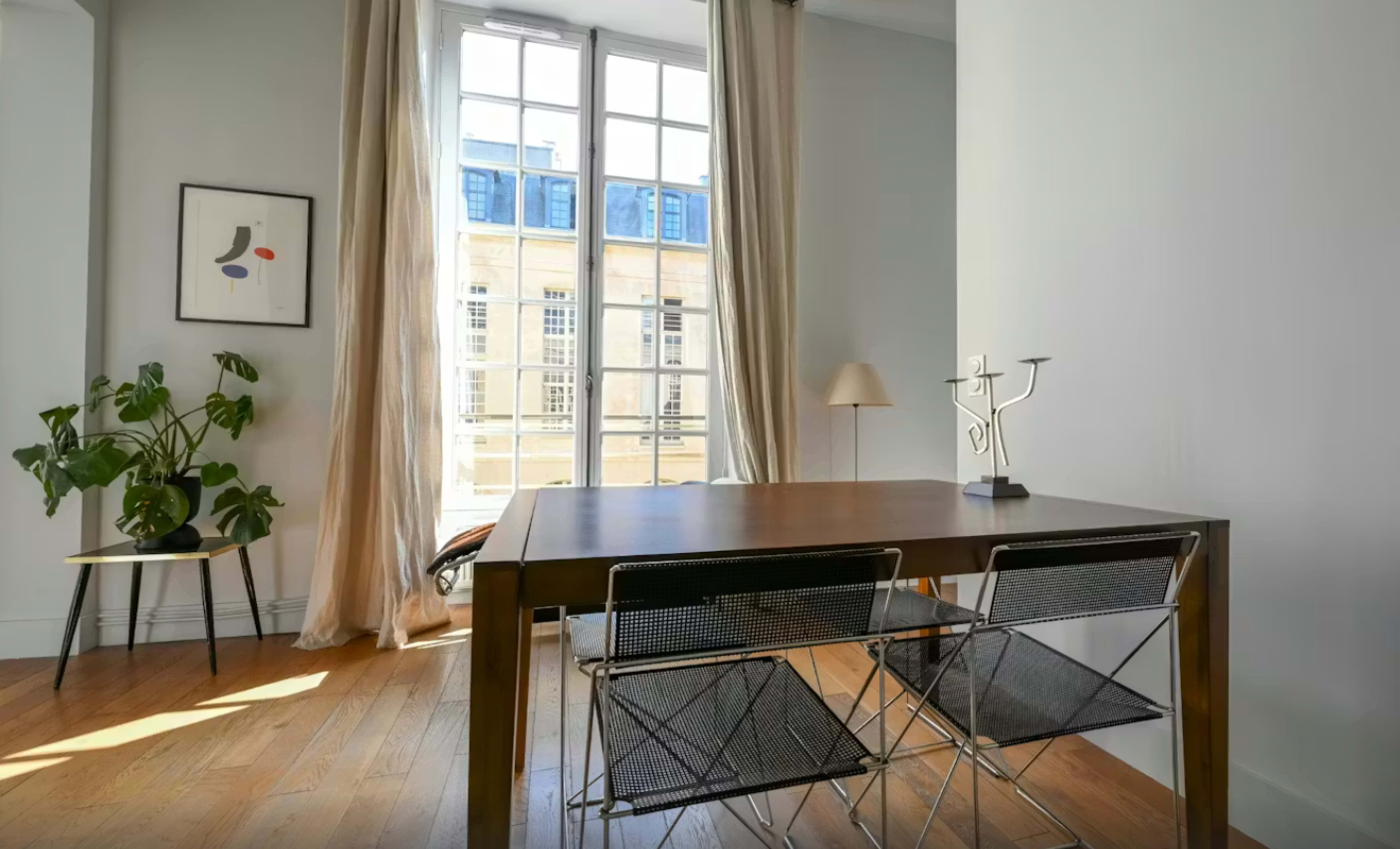 A wooden dining table with two chairs is positioned in a room featuring large windows that allow natural light to enter, complemented by light-colored curtains and a small lamp in the corner.
