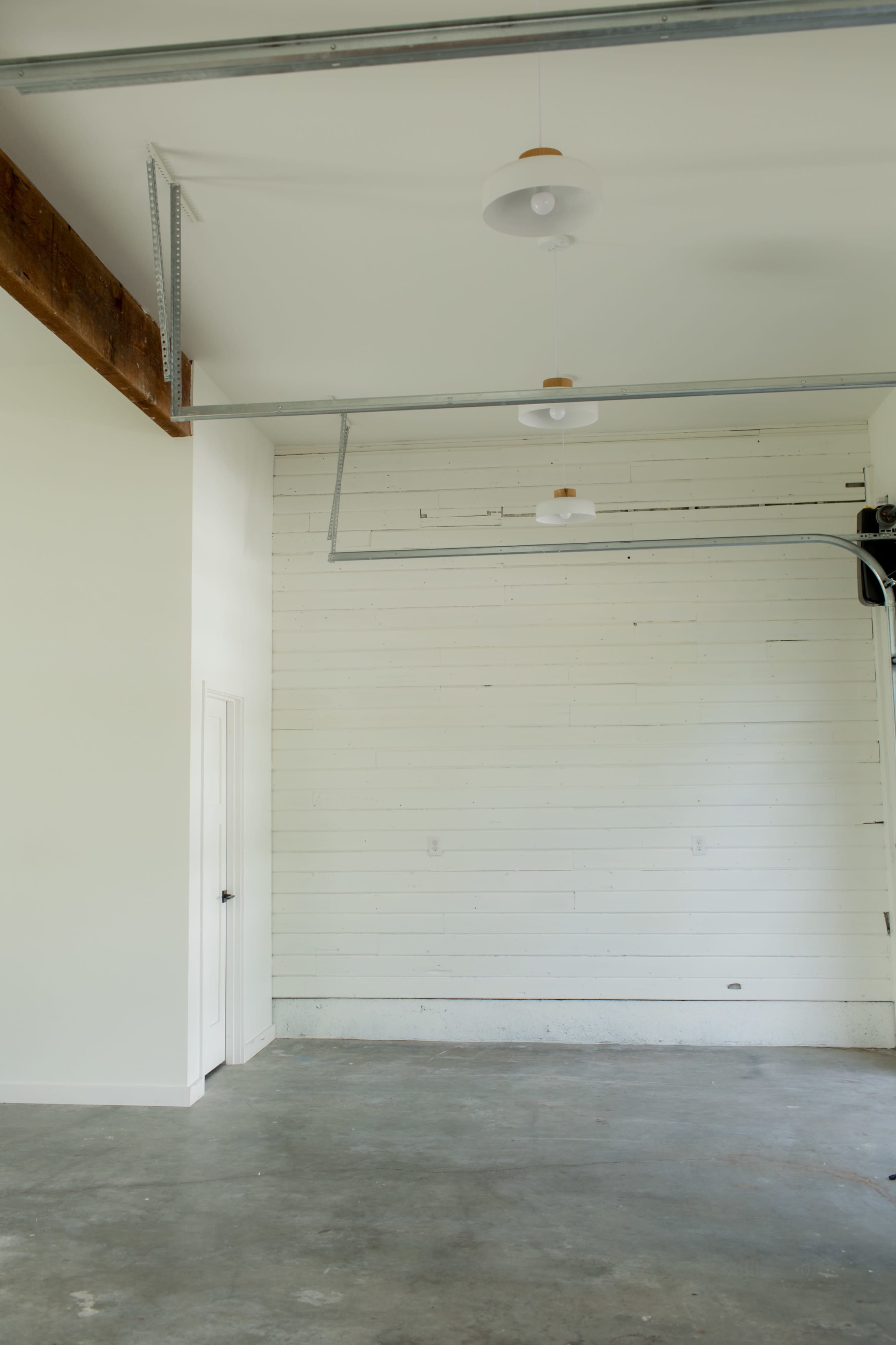 The image shows a spacious, empty interior room with white walls, a ceiling with exposed beams, and a door on the left.