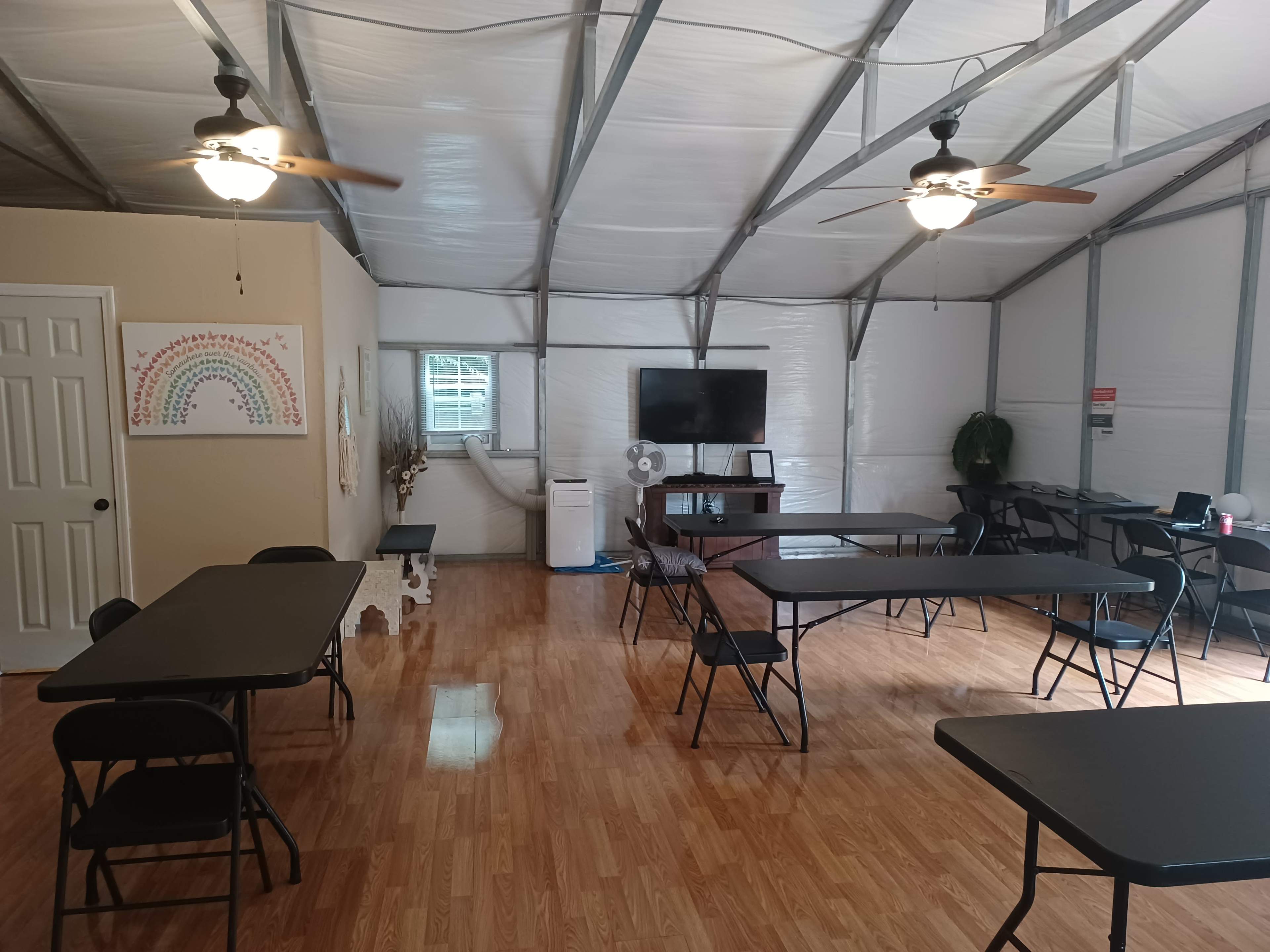 The interior of a spacious room features several black folding tables and chairs, a large television mounted on the wall, and a decorative rainbow painting.