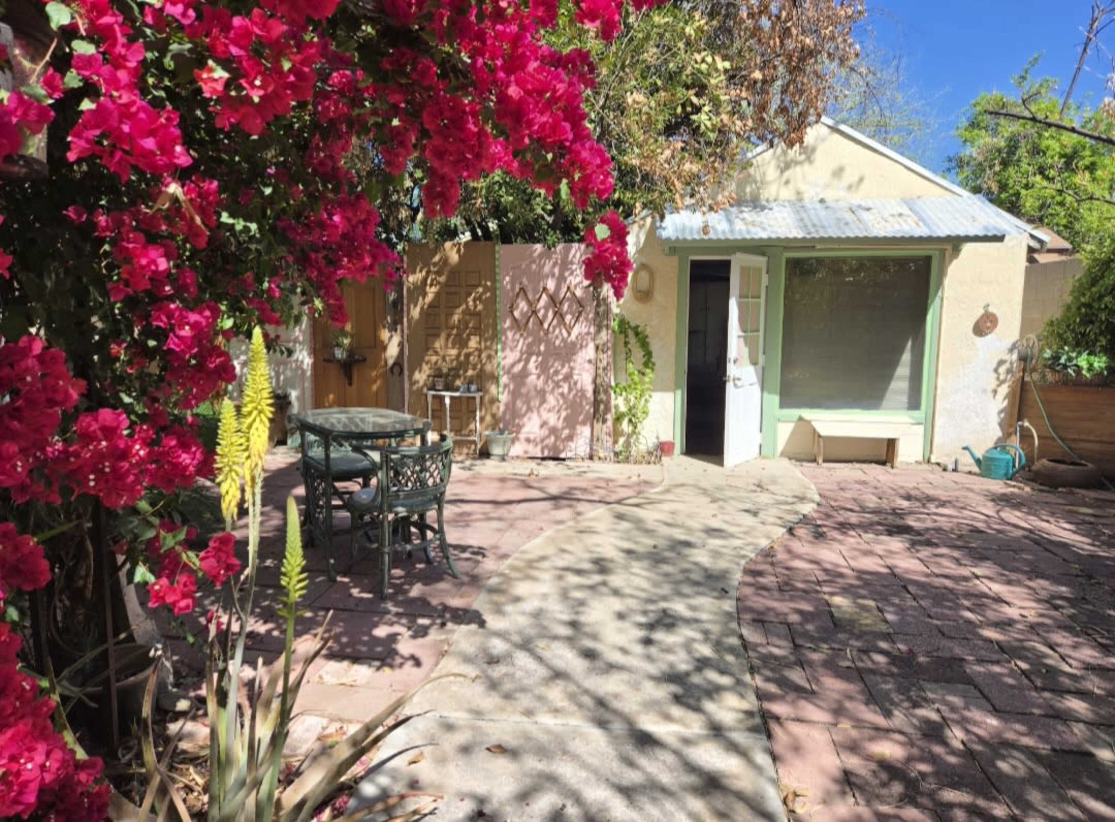 A charming outdoor patio features a stone pathway leading to a small house, framed by vibrant pink bougainvillea and a green border.