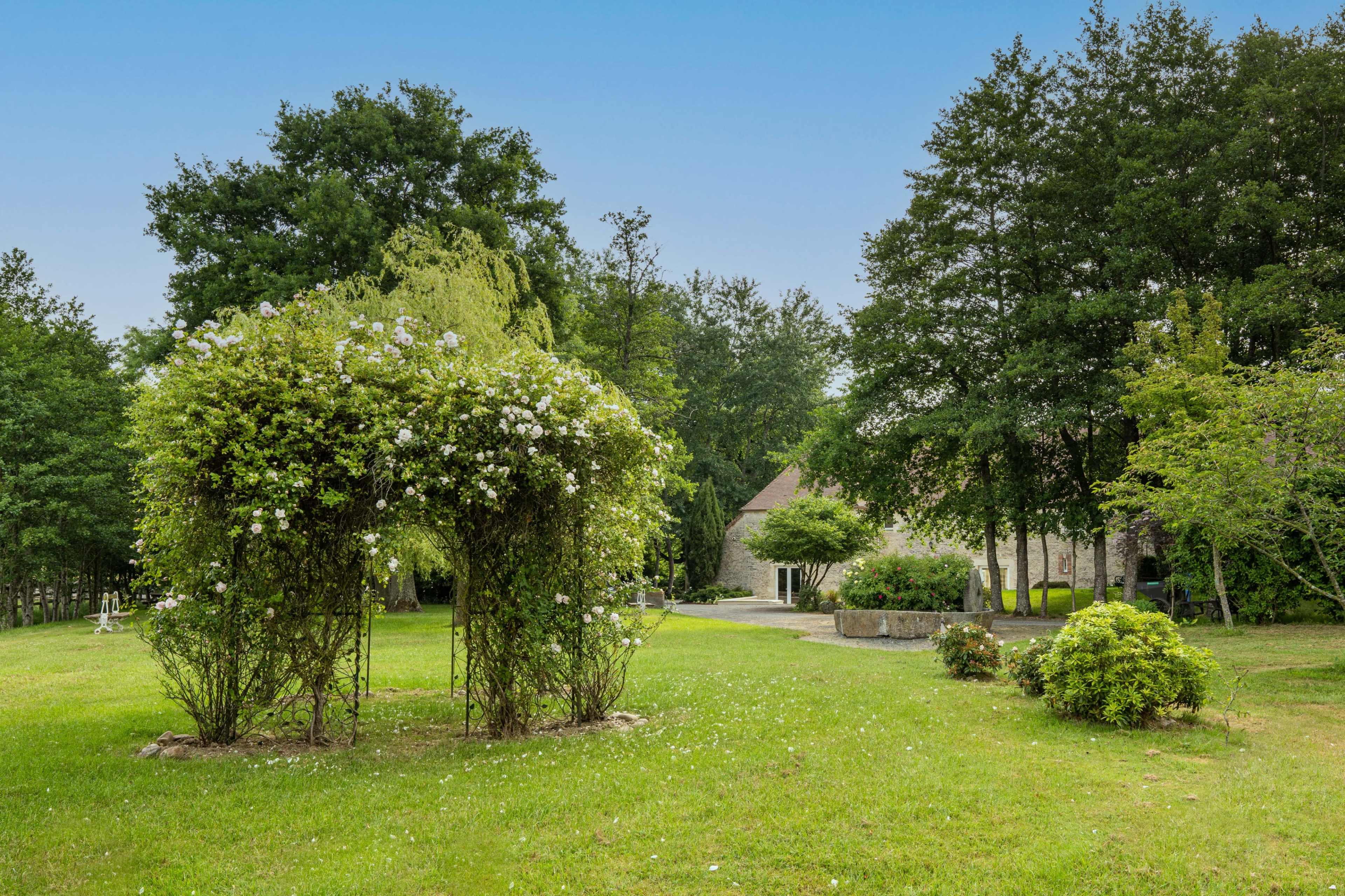 A grassy area features a floral archway leading to a stone building surrounded by trees.