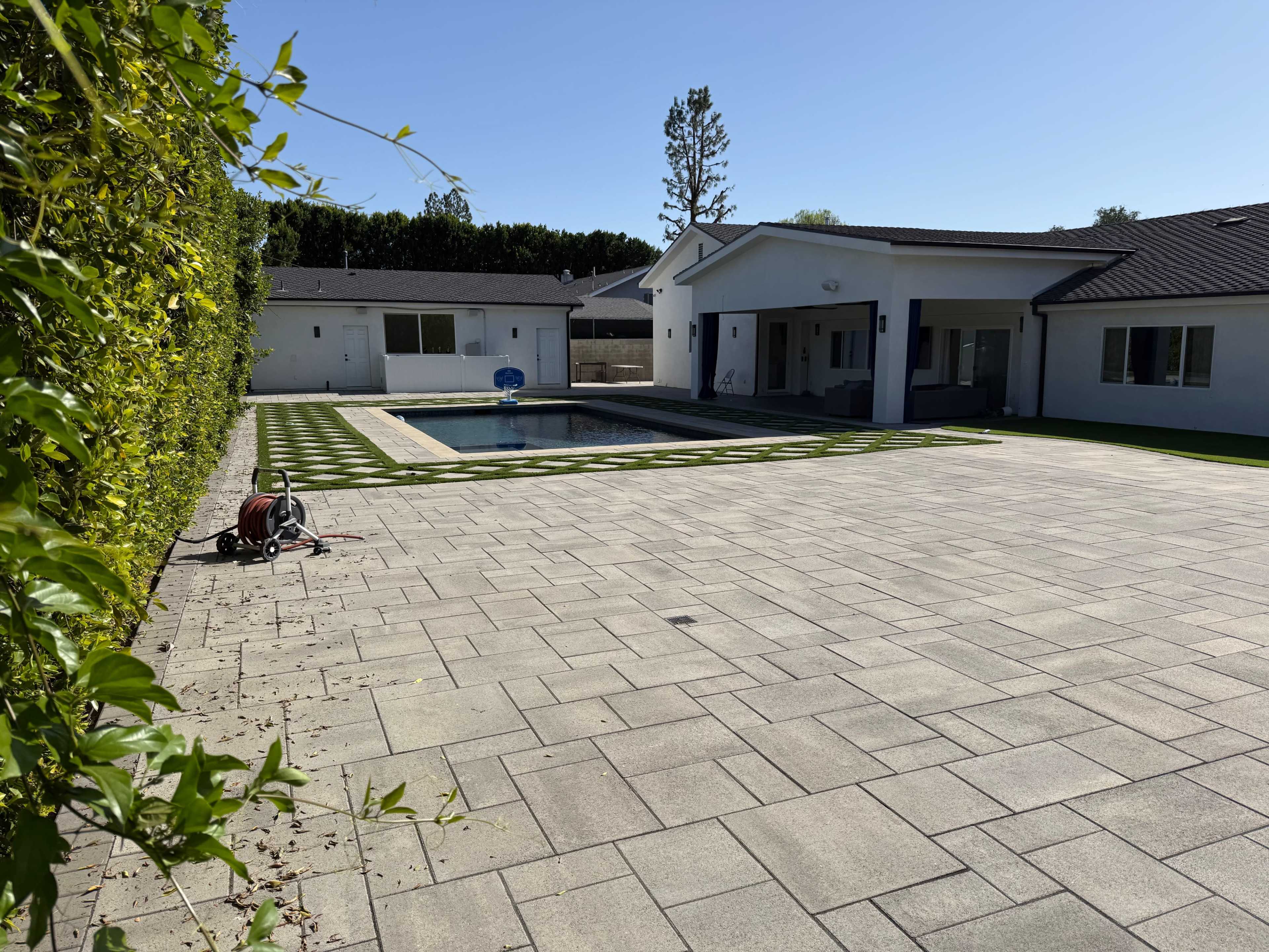 The image shows a spacious outdoor area with a swimming pool, surrounded by a neatly designed patio and a grassy border, set against a backdrop of a modern house.