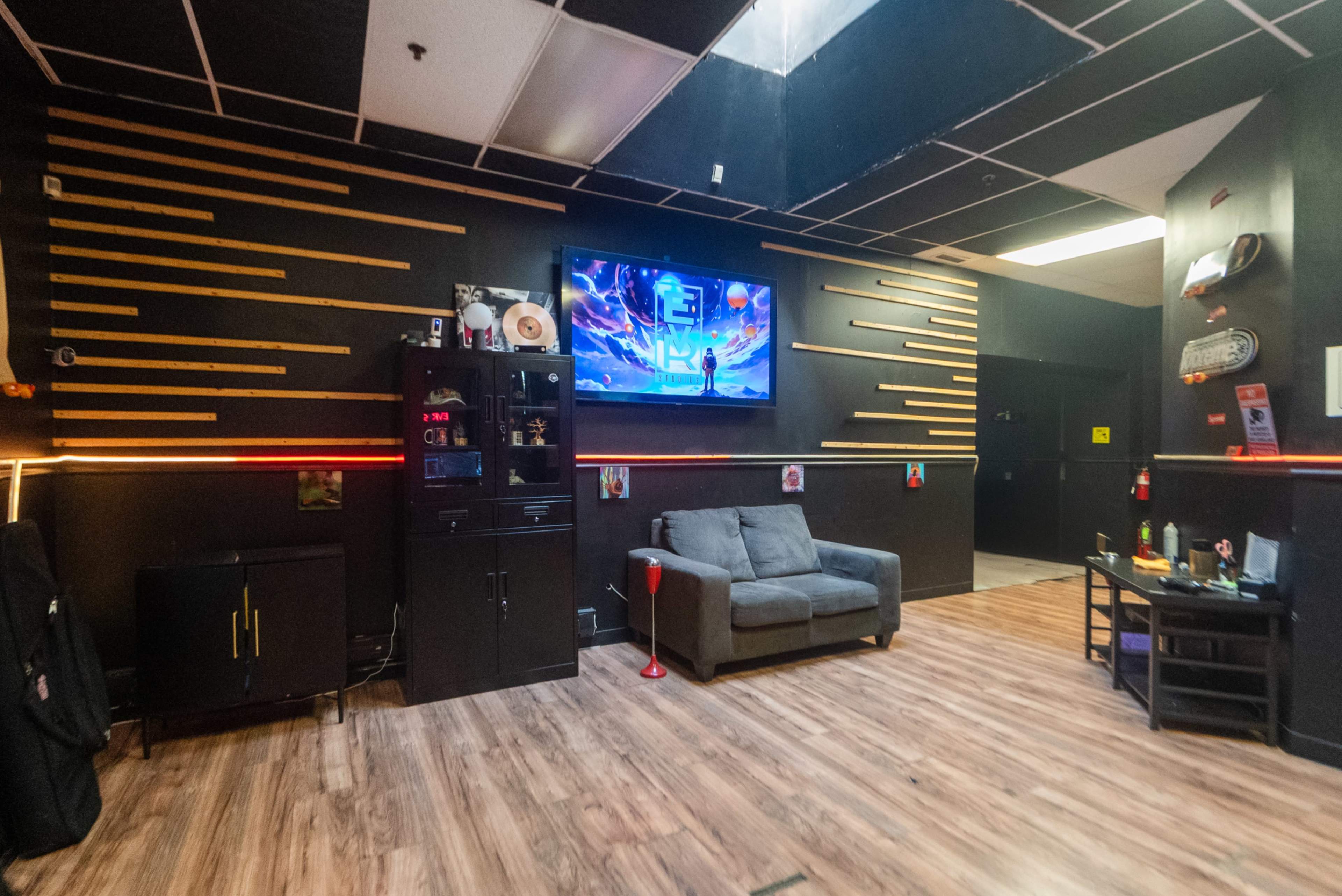 The image shows a modern lounge area with a gray sofa, a wall-mounted television displaying vibrant graphics, and decorative elements made of colored wooden strips against black walls.