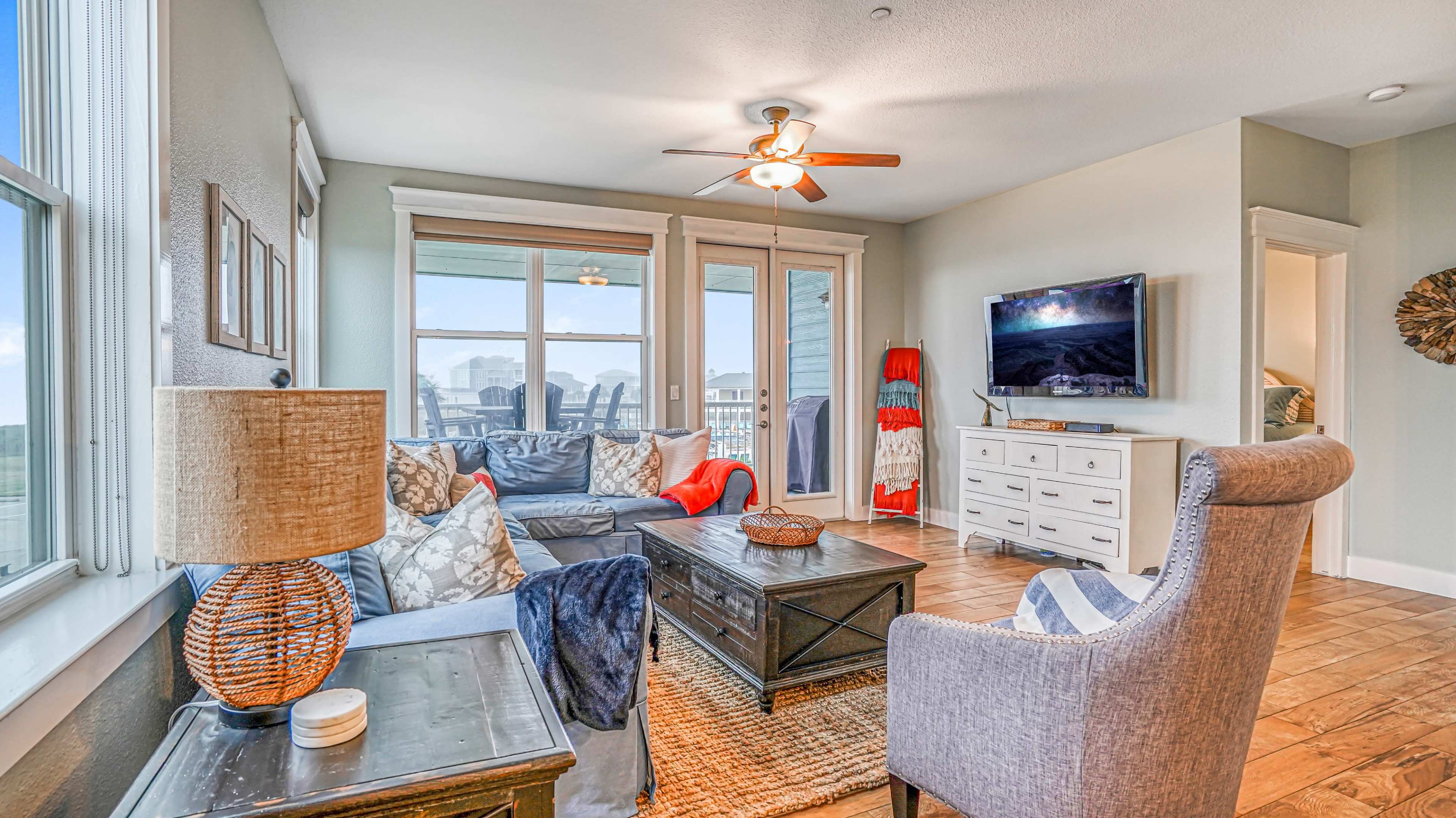 A living room with a ceiling fan, a comfortable seating area, a television on the wall, and large windows letting in natural light.