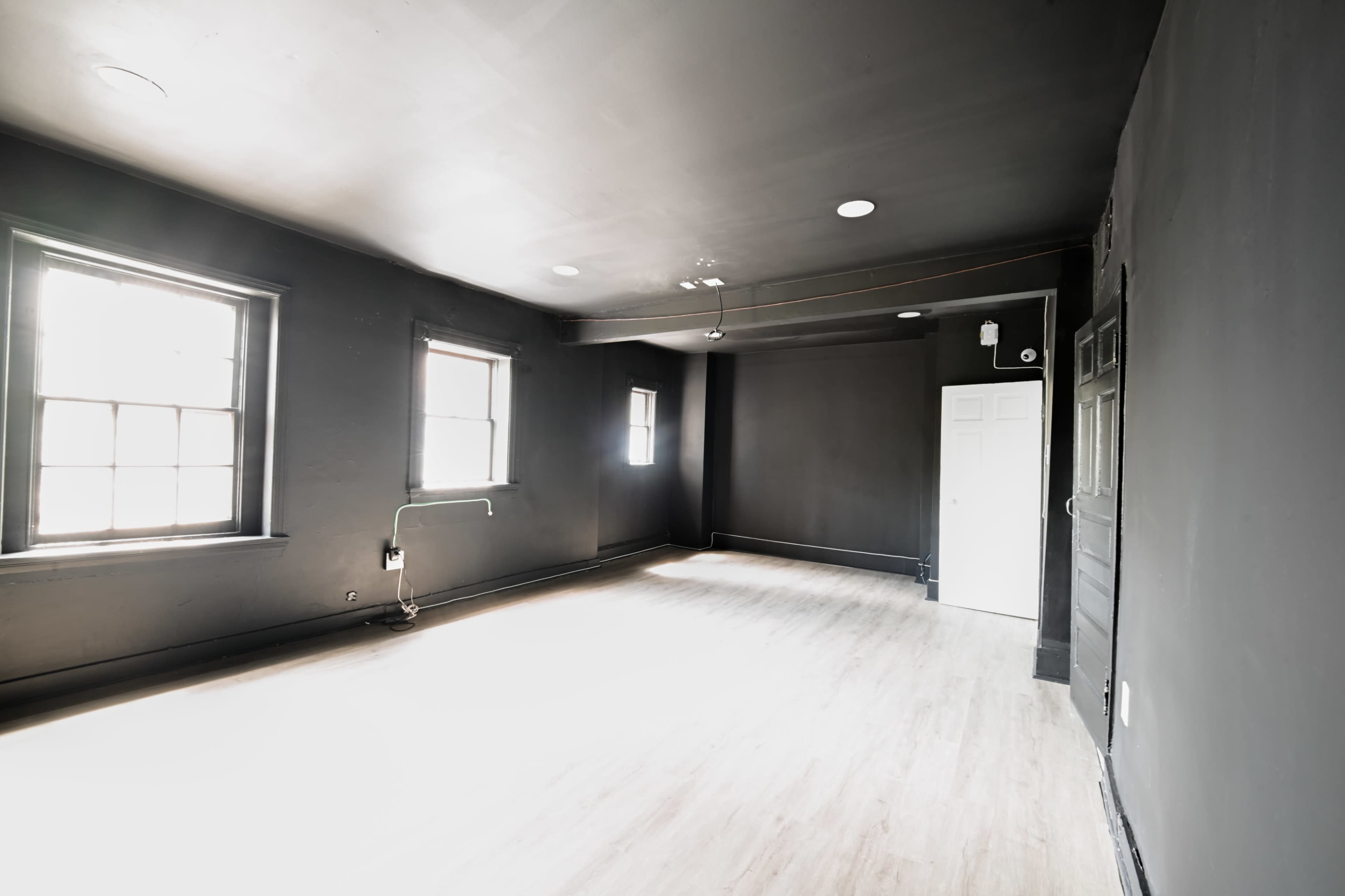 An empty, unfurnished room with black walls and large windows, featuring a light-colored wooden floor and a door at one end.