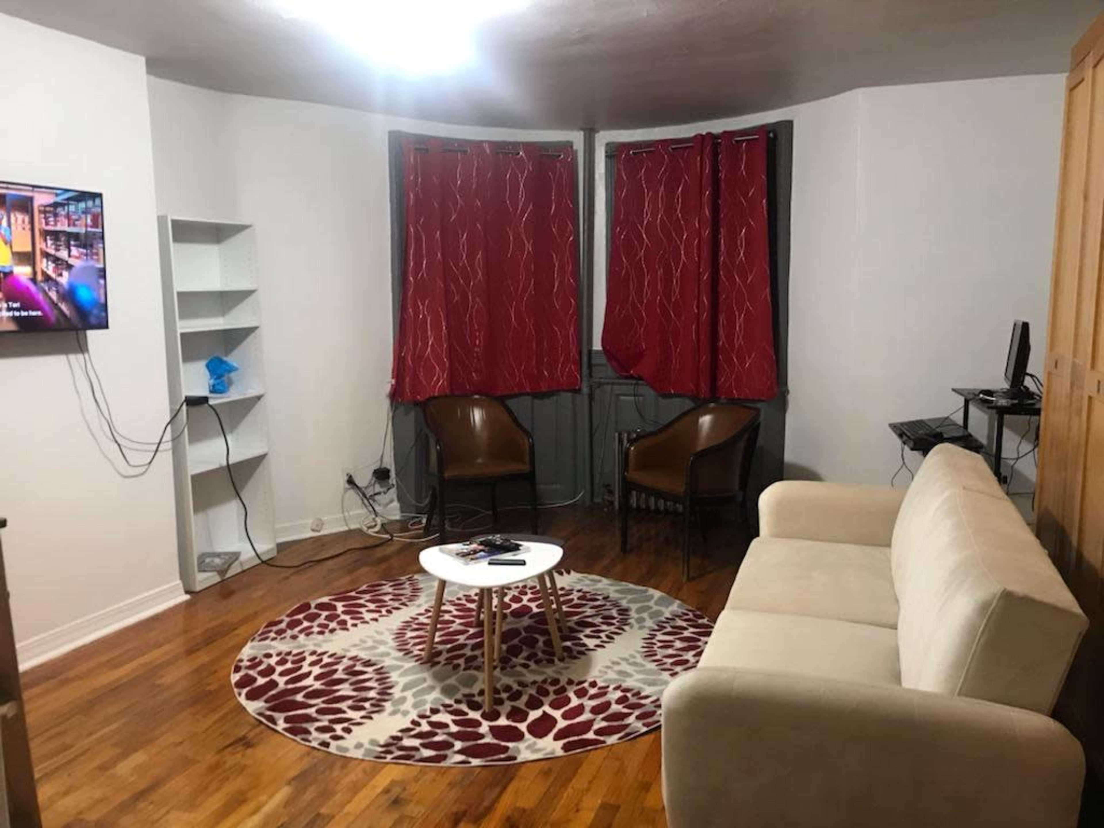 The image shows a small living room with a beige sofa, two brown chairs, a round coffee table, a television on the wall, and red curtains covering the window.