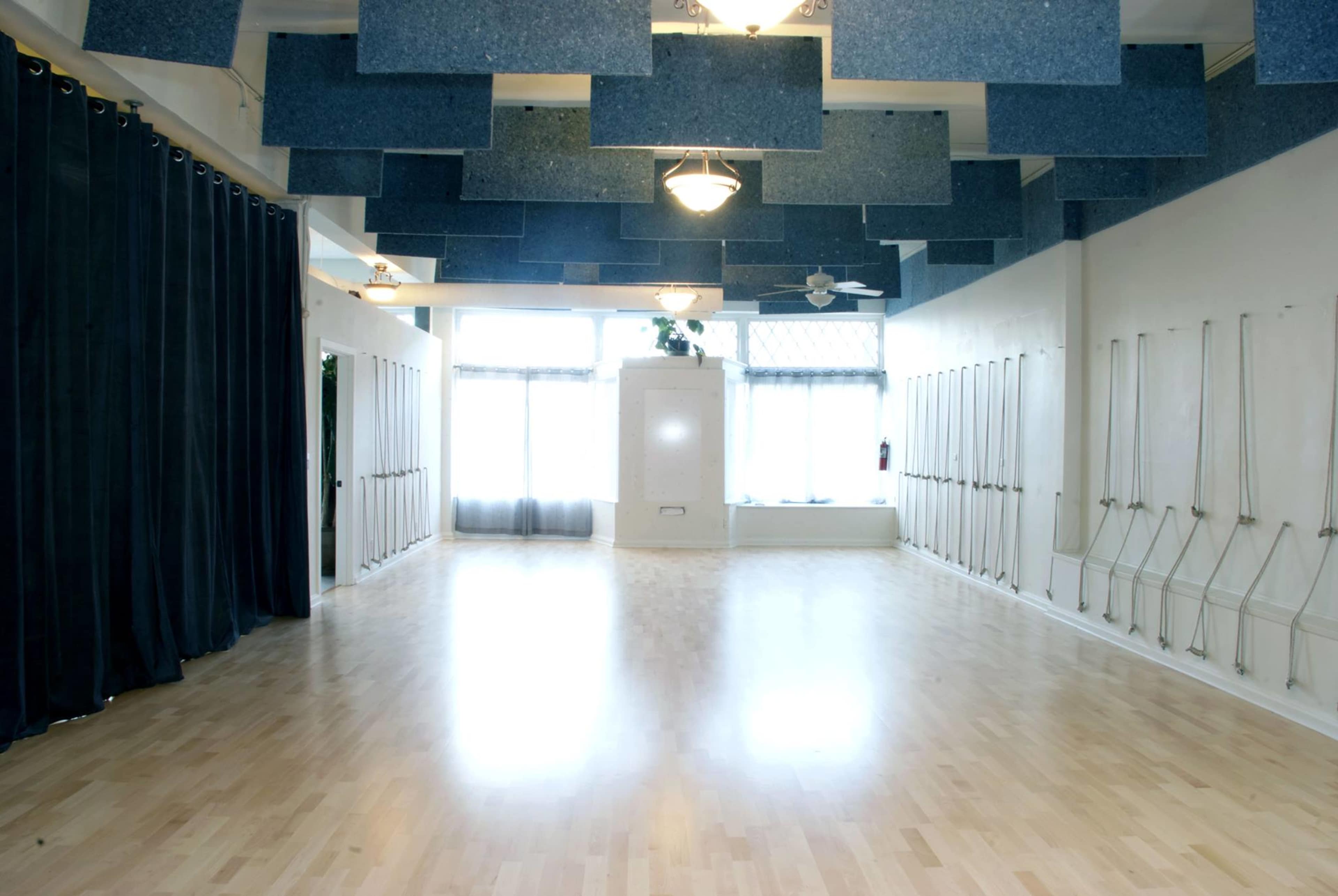 A spacious, well-lit room with large windows, light wooden flooring, and blue acoustic panels hanging from the ceiling.
