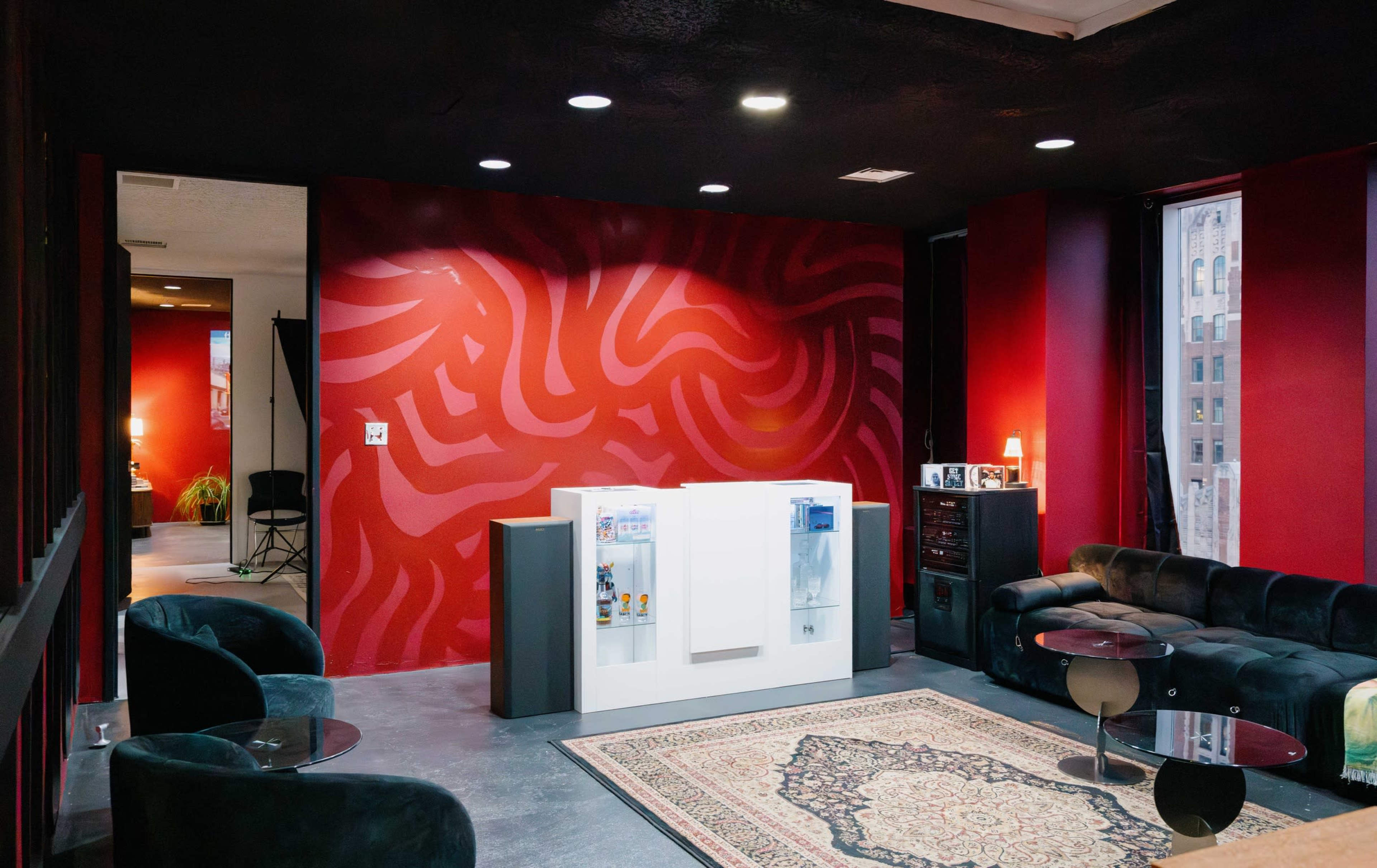 The image shows a modern interior space with red walls, a white refrigerator, and seating arrangements that include a dark couch and coffee table.